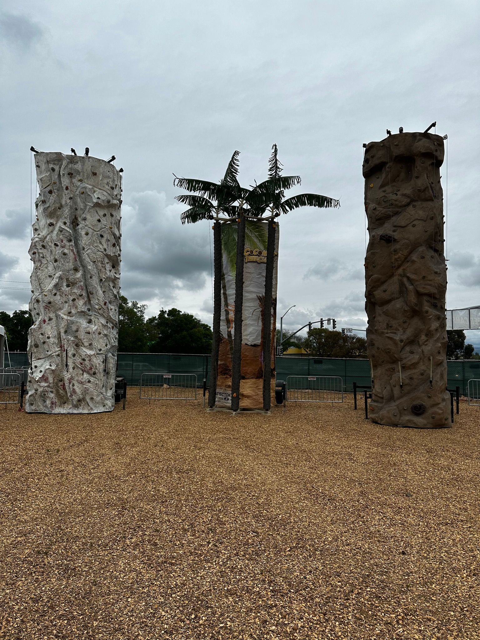 Three vertical rock climbing walls outdoors on a gravel play area under a cloudy sky