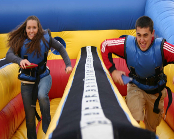 Two people racing side by side on a colorful inflatable slide, smiling and leaning forward