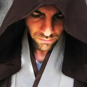 A hooded person in a brown robe looking down, with a focused expression on a dark background.
