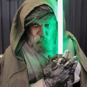 Bearded hooded man holding a glowing green lightsaber with a metallic glove