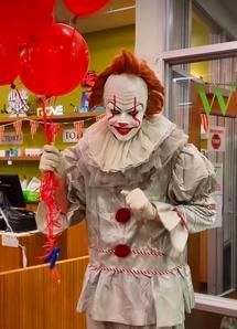 Clown in a white costume with red balloons and a balloon animal, standing indoors and smiling.