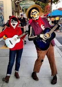 Two masked street performers in costumes play white and blue guitars outdoors.