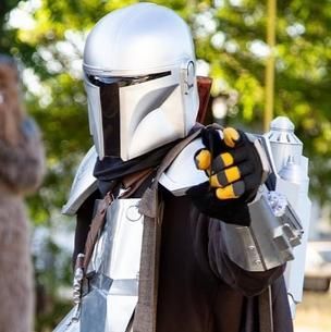 Mandalorian-style armored figure pointing forward, wearing silver helmet and black clothing outdoors