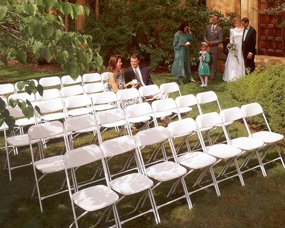 White folding chairs set up on a lawn for an outdoor wedding, with a few guests standing nearby