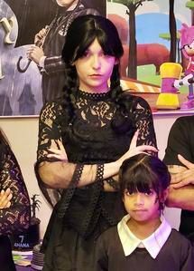 Two people pose indoors; one in a black lace dress stands with arms crossed, another stands in front wearing a white collar.