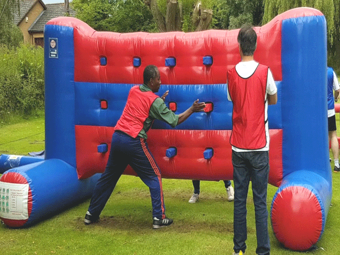 Two people playing a red-and-blue inflatable game outdoors, aiming at targets on a padded wall