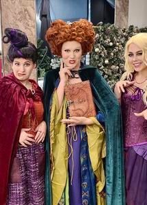Three people in colorful witch costumes pose together, with dramatic makeup and Halloween-style props.