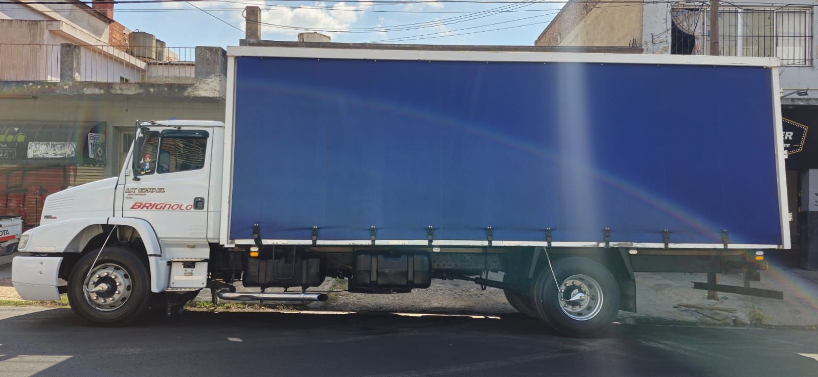 Un camión blanco con un gran contenedor de carga azul estacionado en una calle frente a unos edificios.
