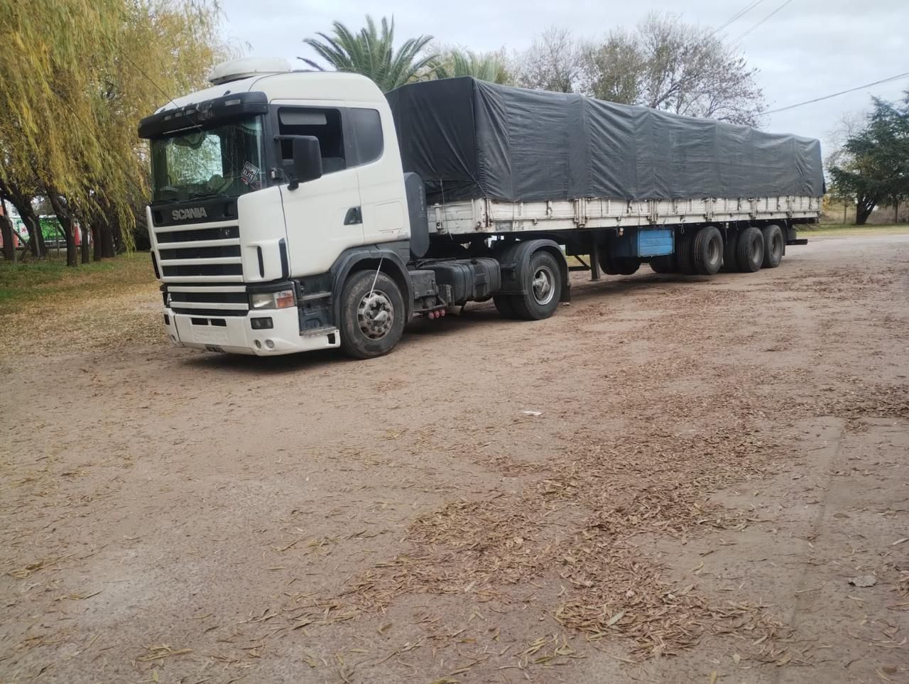 Camión de carga blanco con remolque cubierto estacionado en un camino de tierra cerca de árboles.