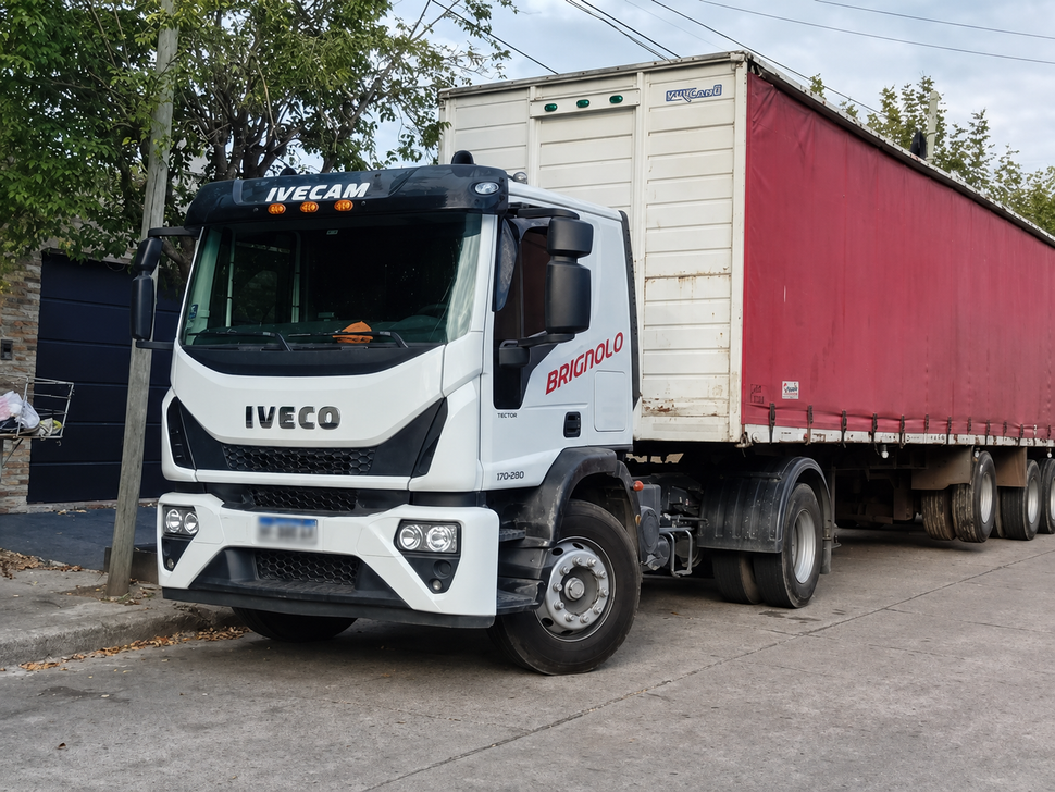 Un camión semirremolque Iveco blanco arrastrando un remolque rojo en la cuneta, cerca de árboles y edificios.