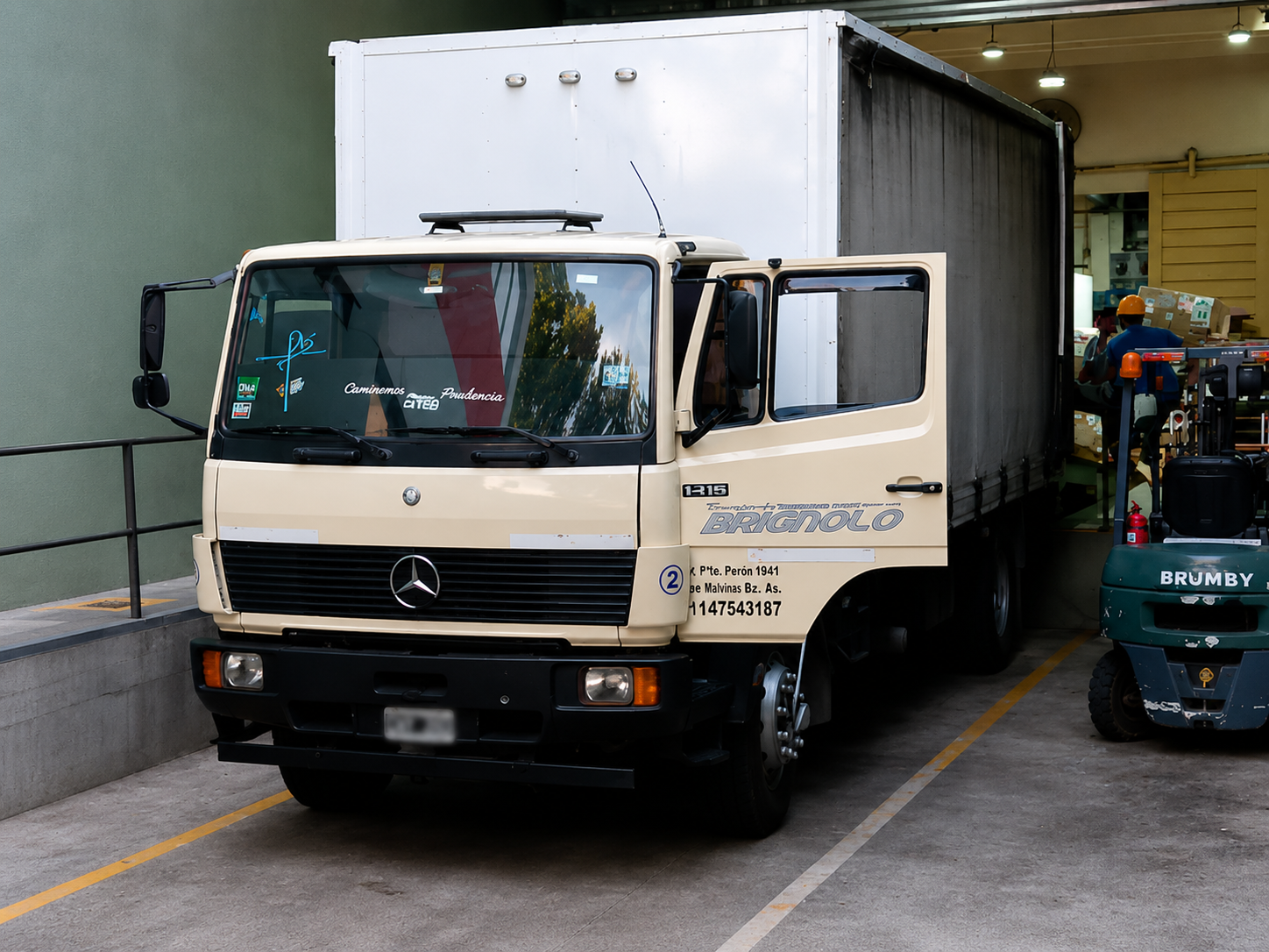 Un camión Mercedes de color beige estacionado en un taller, con una carretilla elevadora verde al lado.