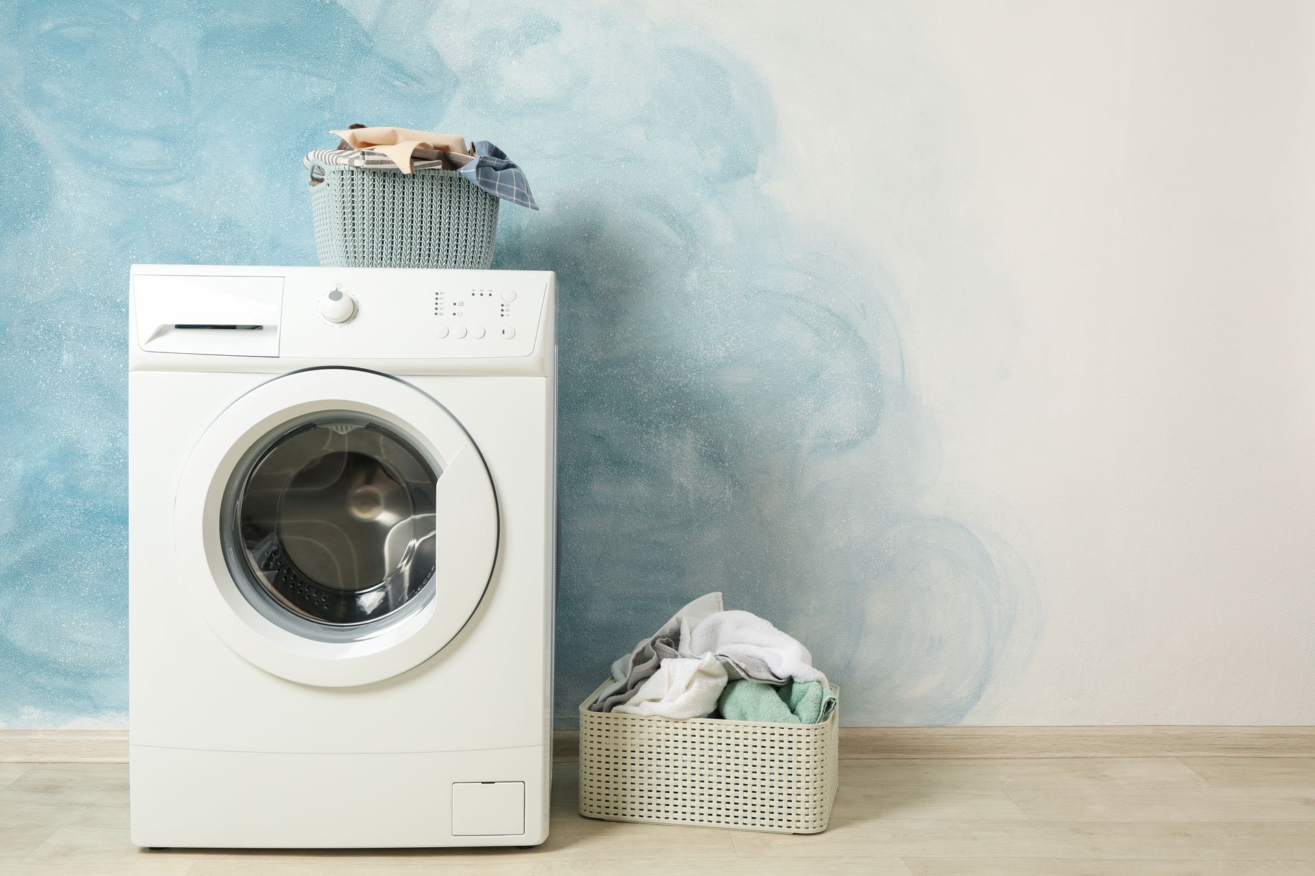 Washing machine with laundry basket on top and a basket of clothes beside it against a blue wall. Washing machine with laundry basket on top and a basket of clothes beside it against a blue wall.