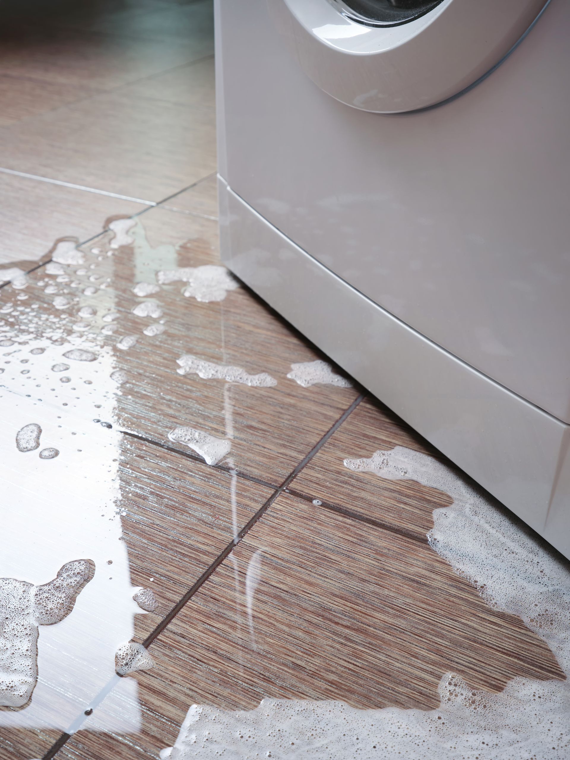 Washing machine leaking water onto a tile floor. White machine, brown and beige tiles. Washing machine leaking water onto a tile floor. White machine, brown and beige tiles.