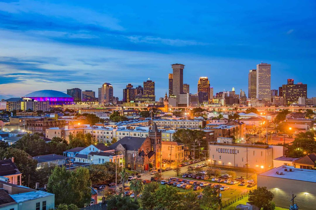 City skyline at dusk with glowing lights and a purple-lit stadium under a blue evening sky