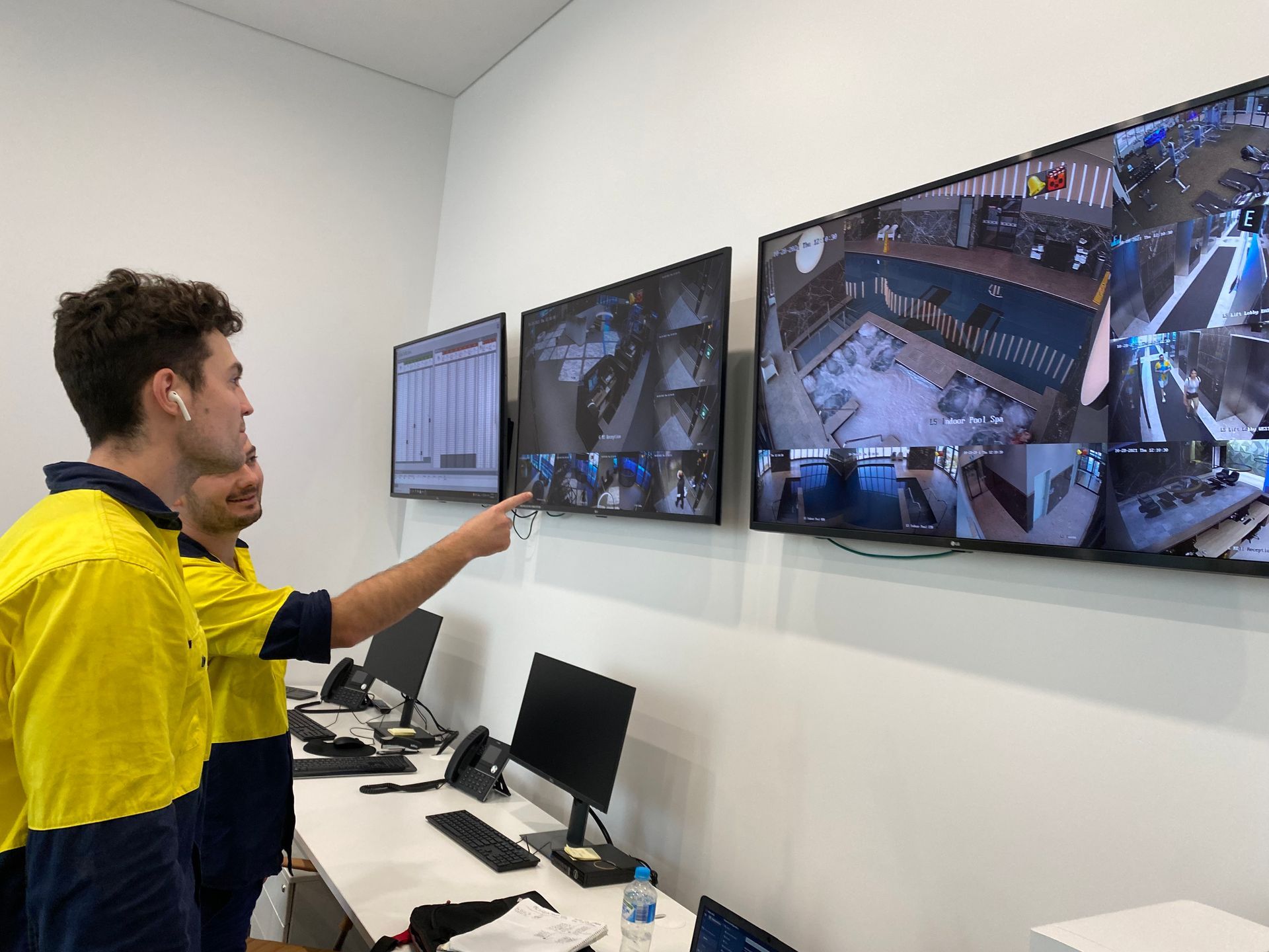 Technician in hi-vis pointing at multiple CCTV monitoring screens showing live camera feeds.