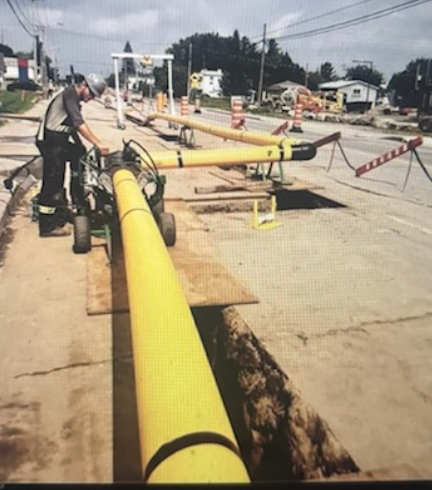 Man operating machinery, installing yellow pipes in a road trench. Sidewalk, buildings, and construction signs visible.