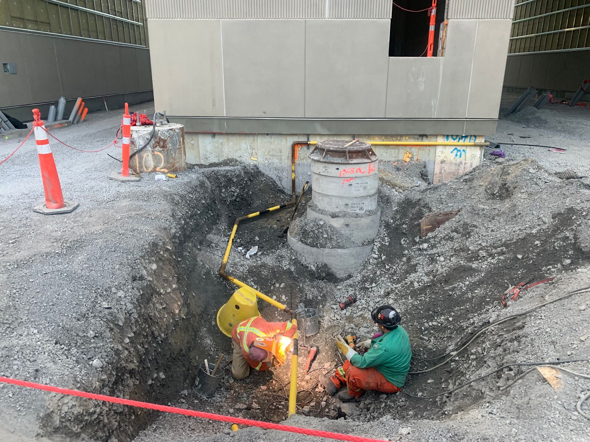 Two construction workers welding a pipe in a trench, near a concrete structure and road cones.