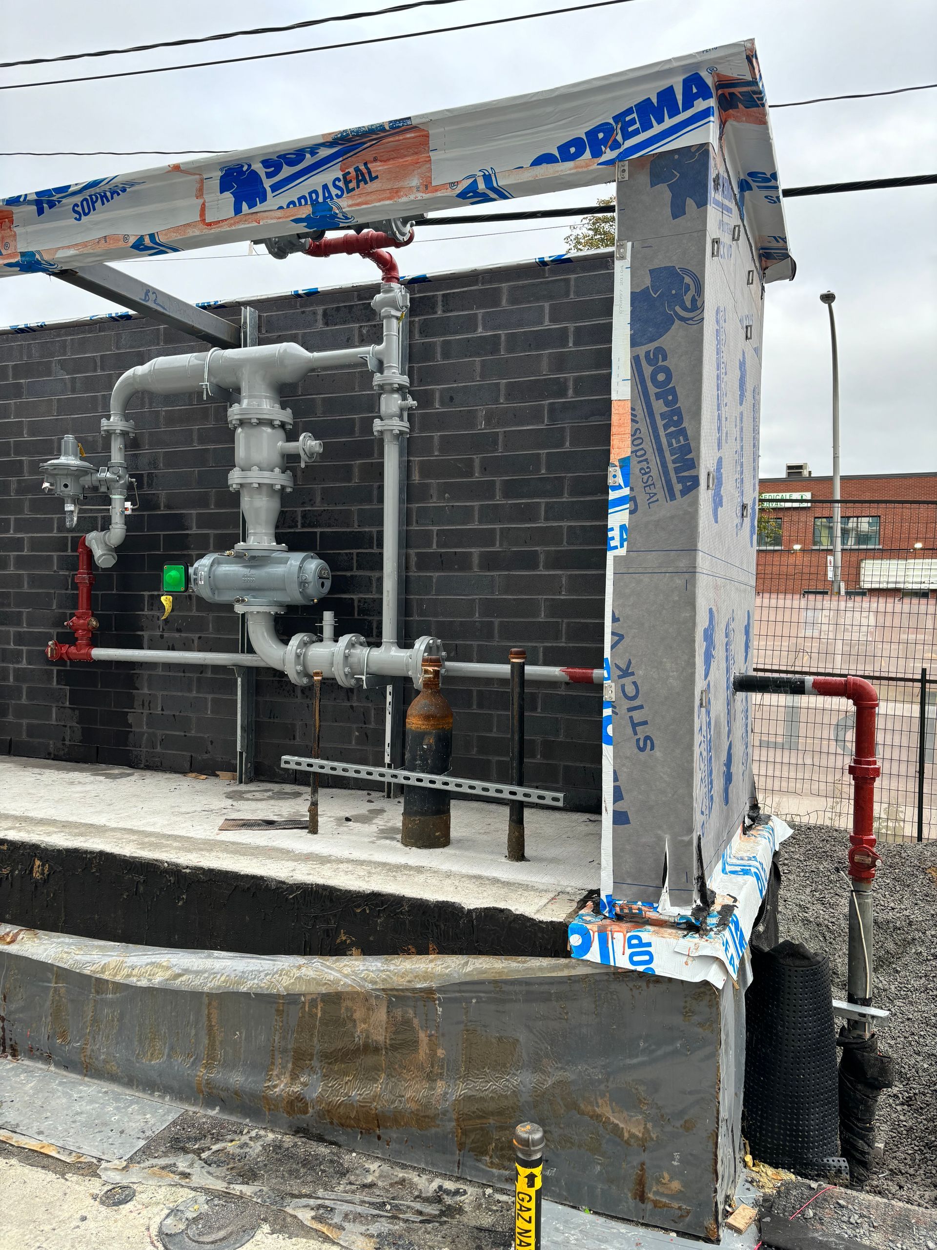 Gas pipeline equipment mounted on a brick wall, with red and silver pipes. Concrete foundation in foreground.