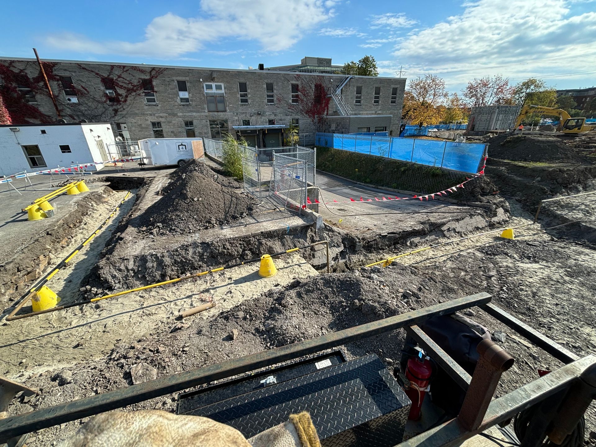 Construction site with excavated areas, a brick building in the background, and blue protective fencing.
