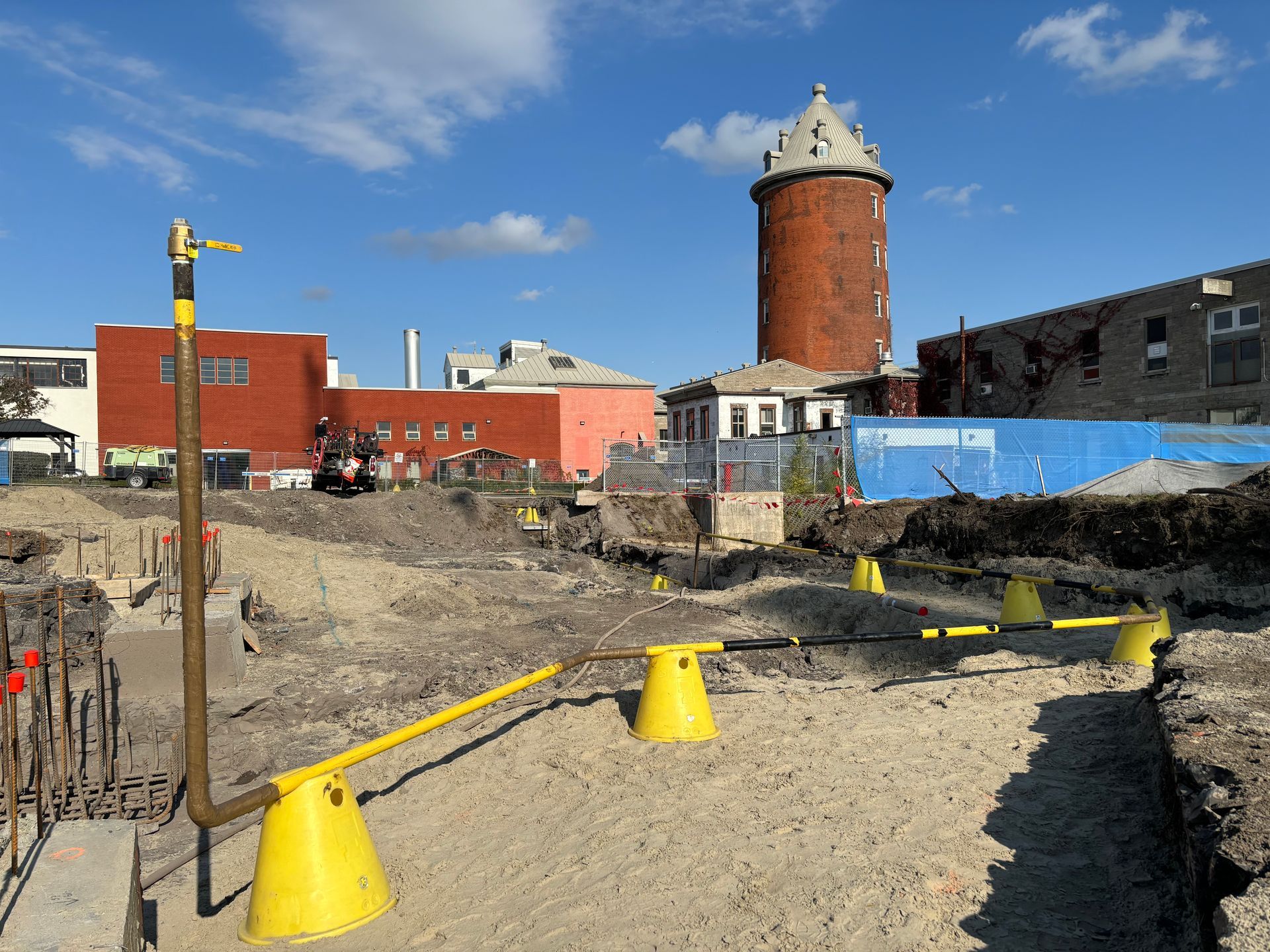 Construction site with yellow safety cones, a red brick tower, and buildings under a blue sky.