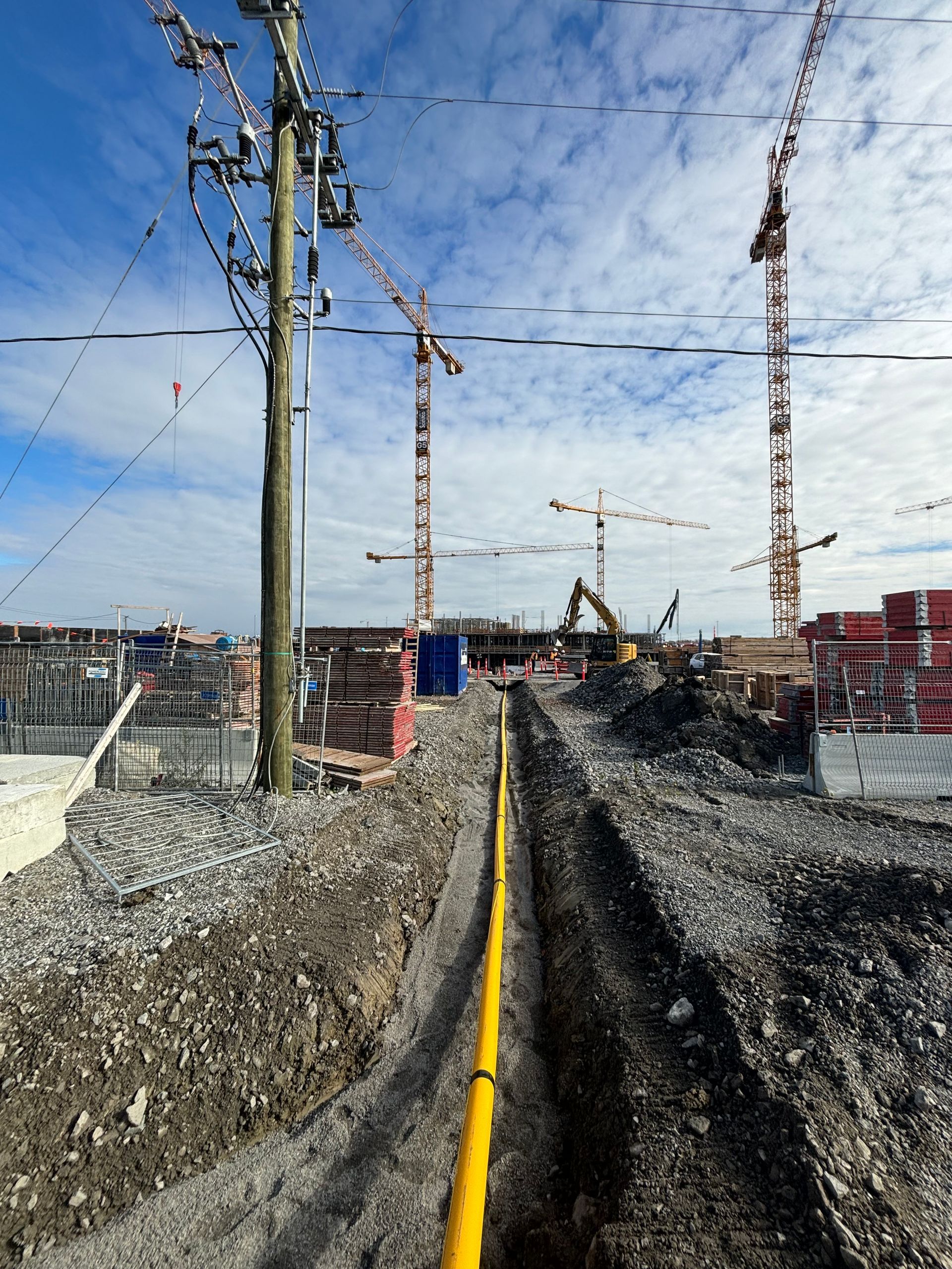 A construction site with a trench, yellow pipe, power lines, and cranes against a cloudy sky.