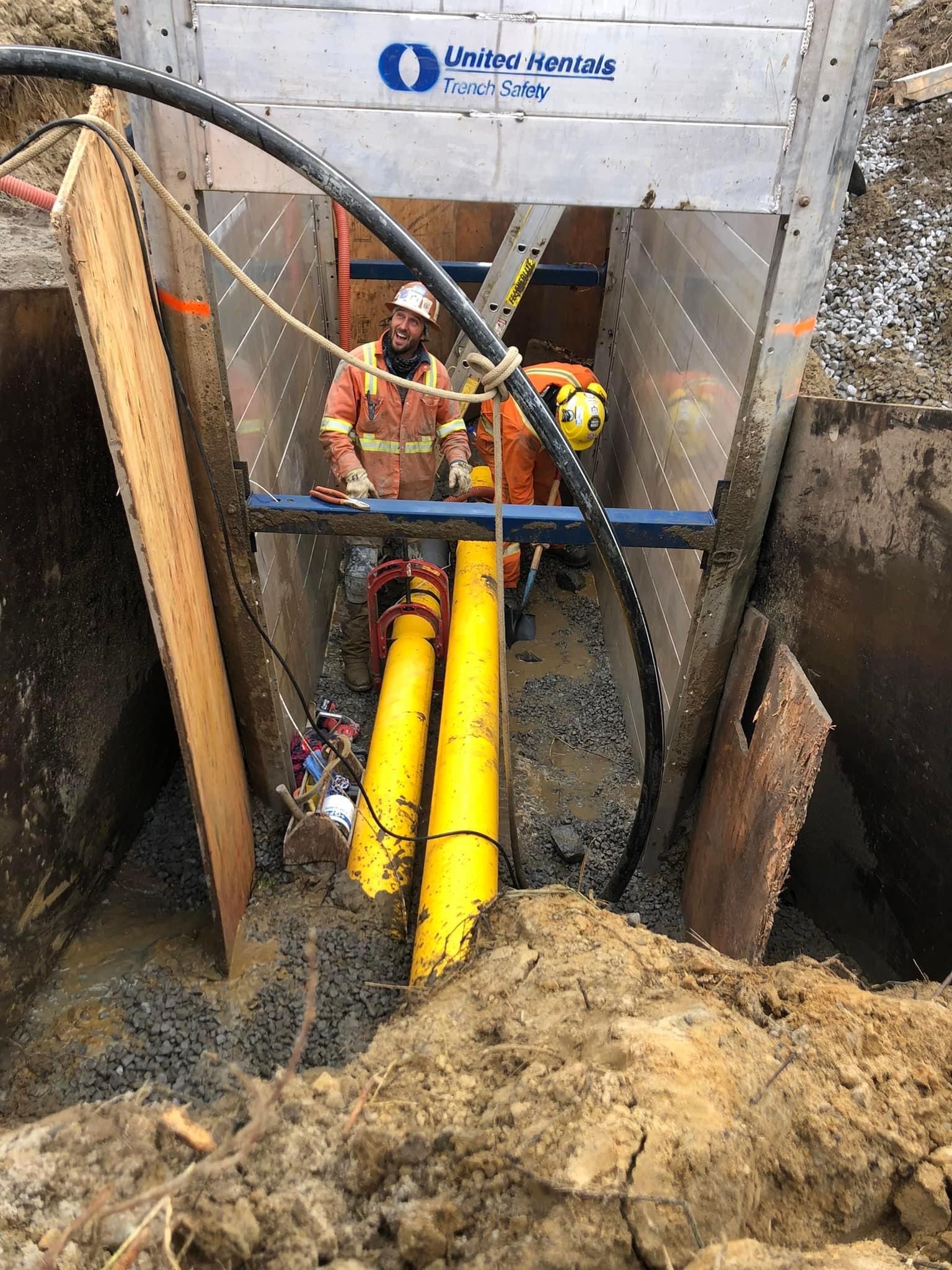 Two workers in a trench, installing yellow pipes. Aluminum trench box provides safety.