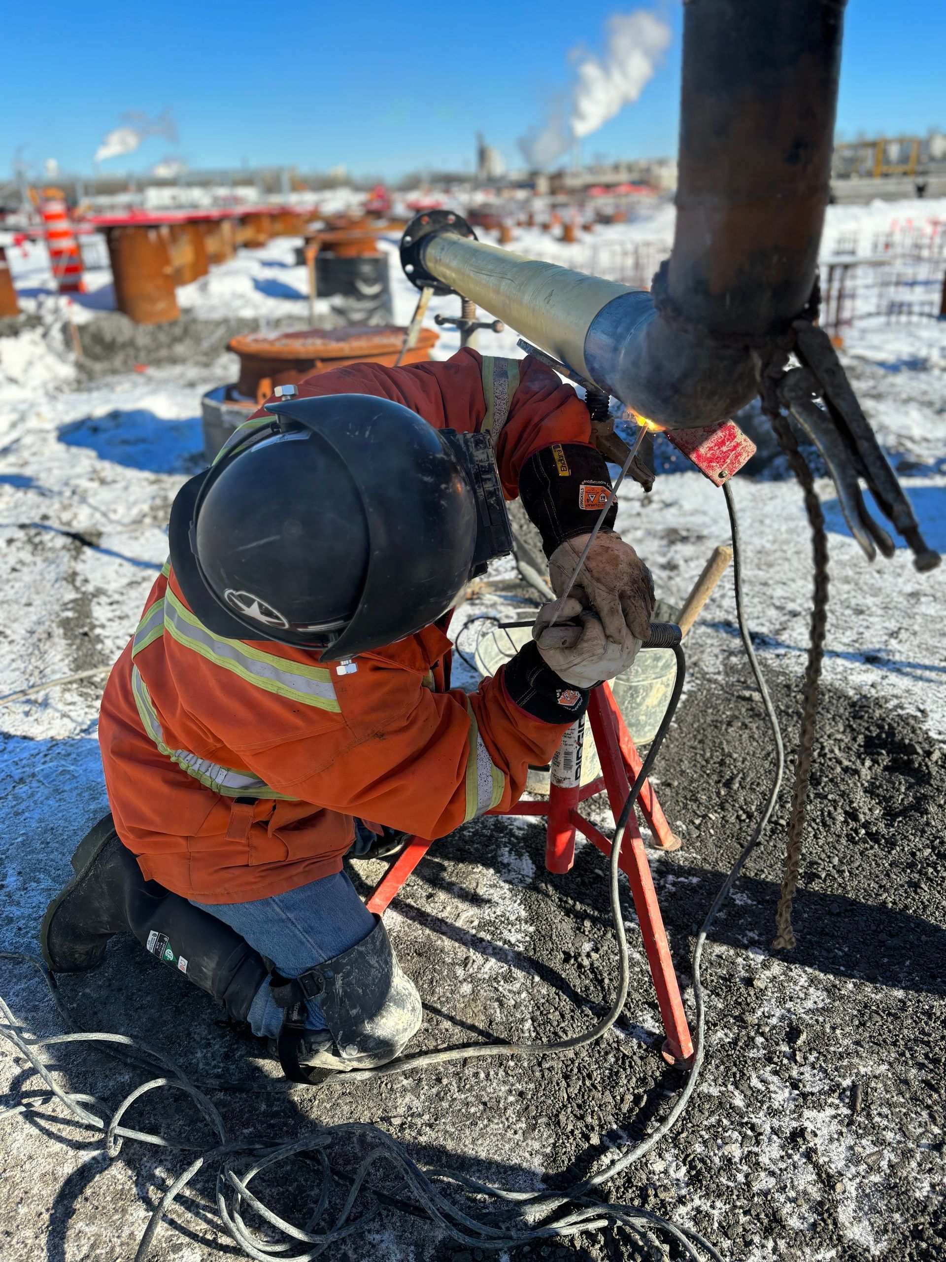 A worker in orange safety gear welds a pipe outdoors in a snowy construction site.