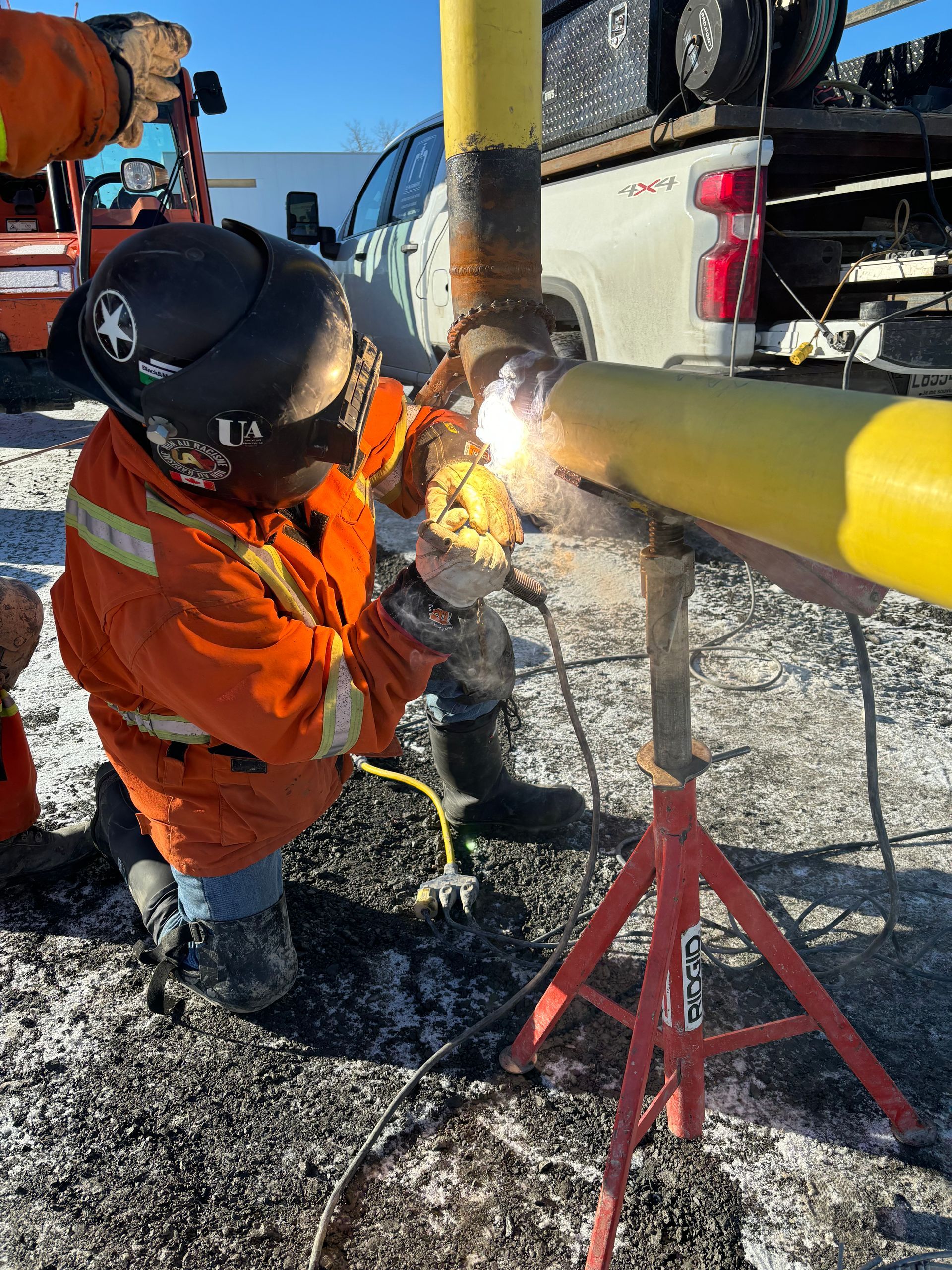 Welder in orange suit welding yellow pipe outdoors.