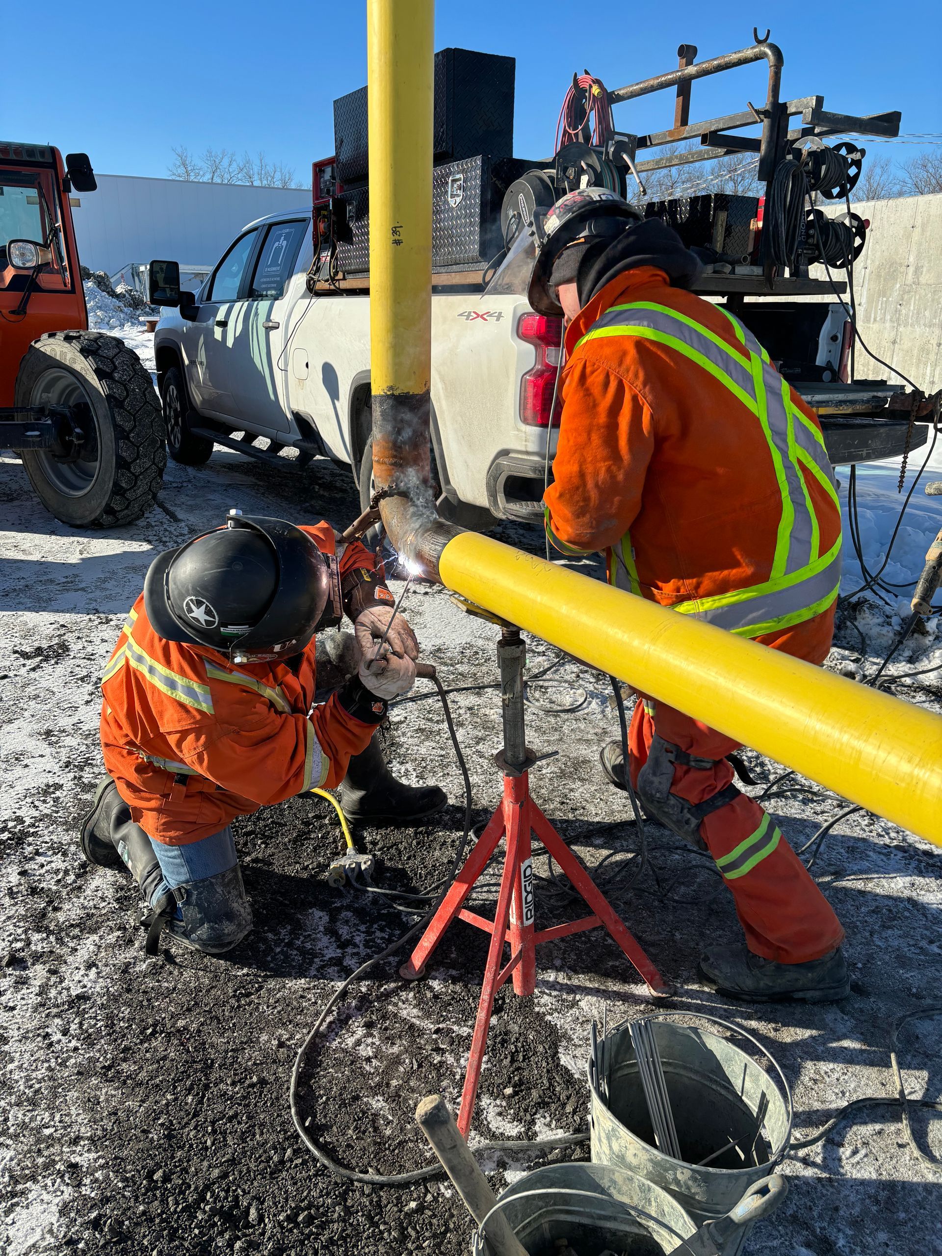 Two workers in orange suits welding a yellow pipe in a snowy outdoor setting.