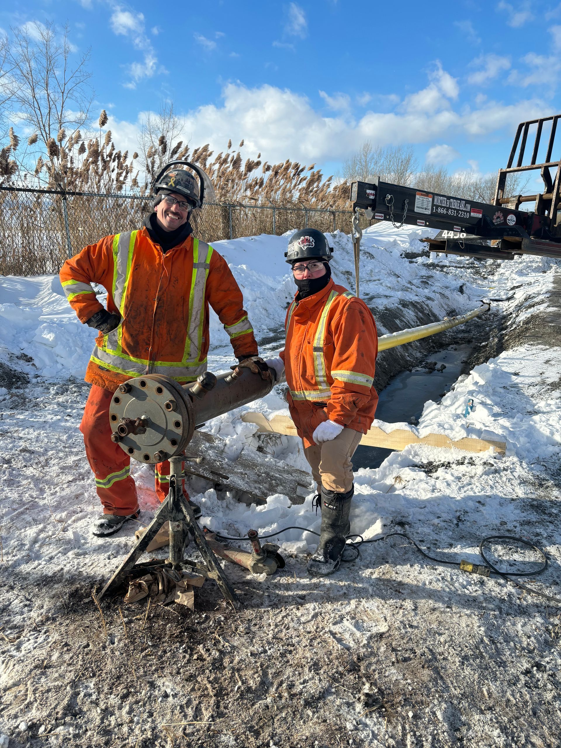 Two people in orange work suits stand near a damaged piece of machinery on a snowy day.