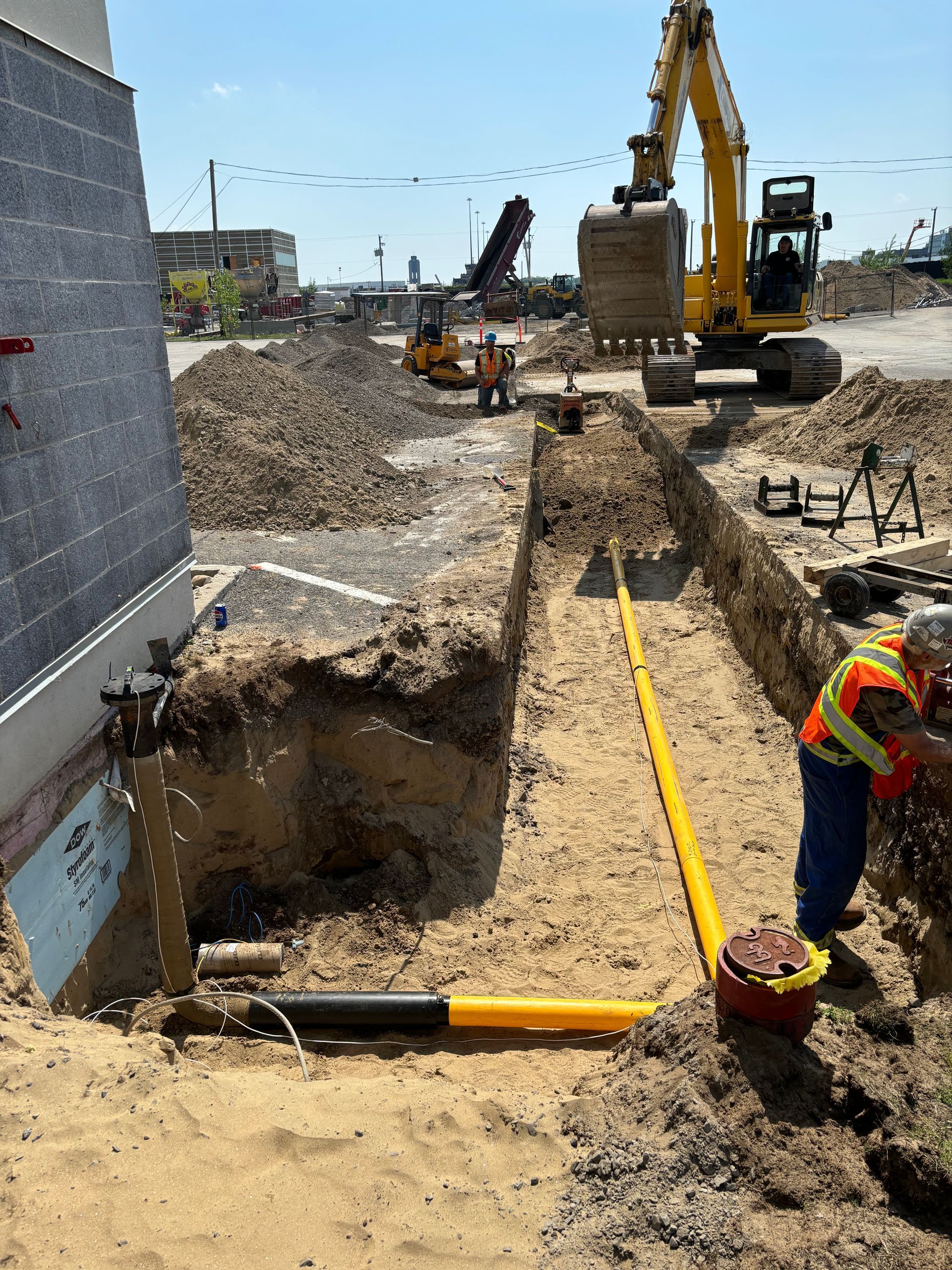 Construction site: workers installing yellow gas lines in trench with excavator and building.