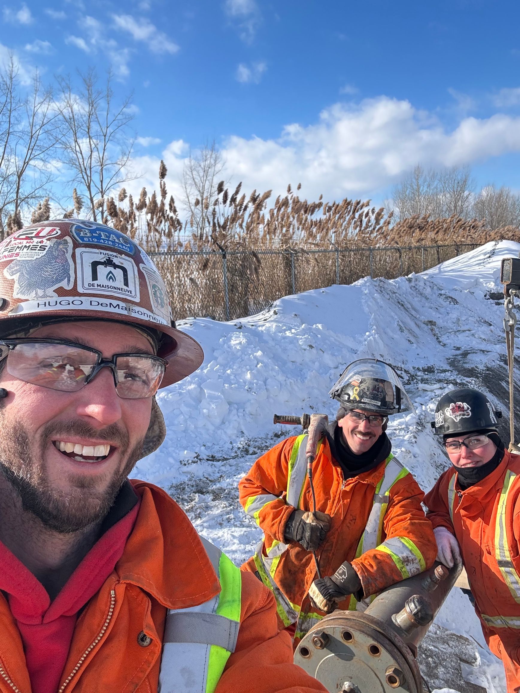 Three workers in orange safety gear and helmets smile outside in the snow.