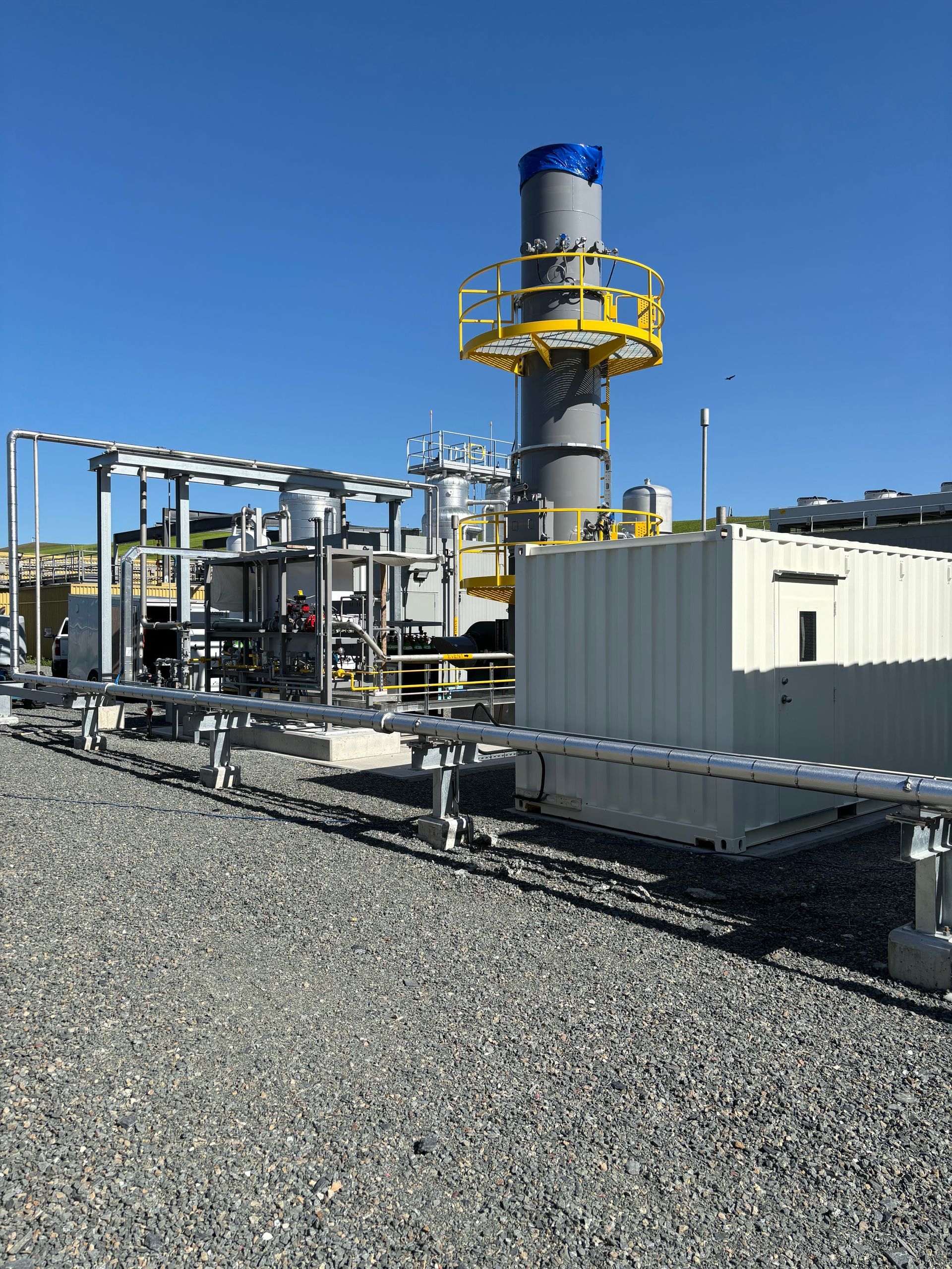 Industrial facility with a tall smokestack and pipes, set on a gravel lot under a blue sky.