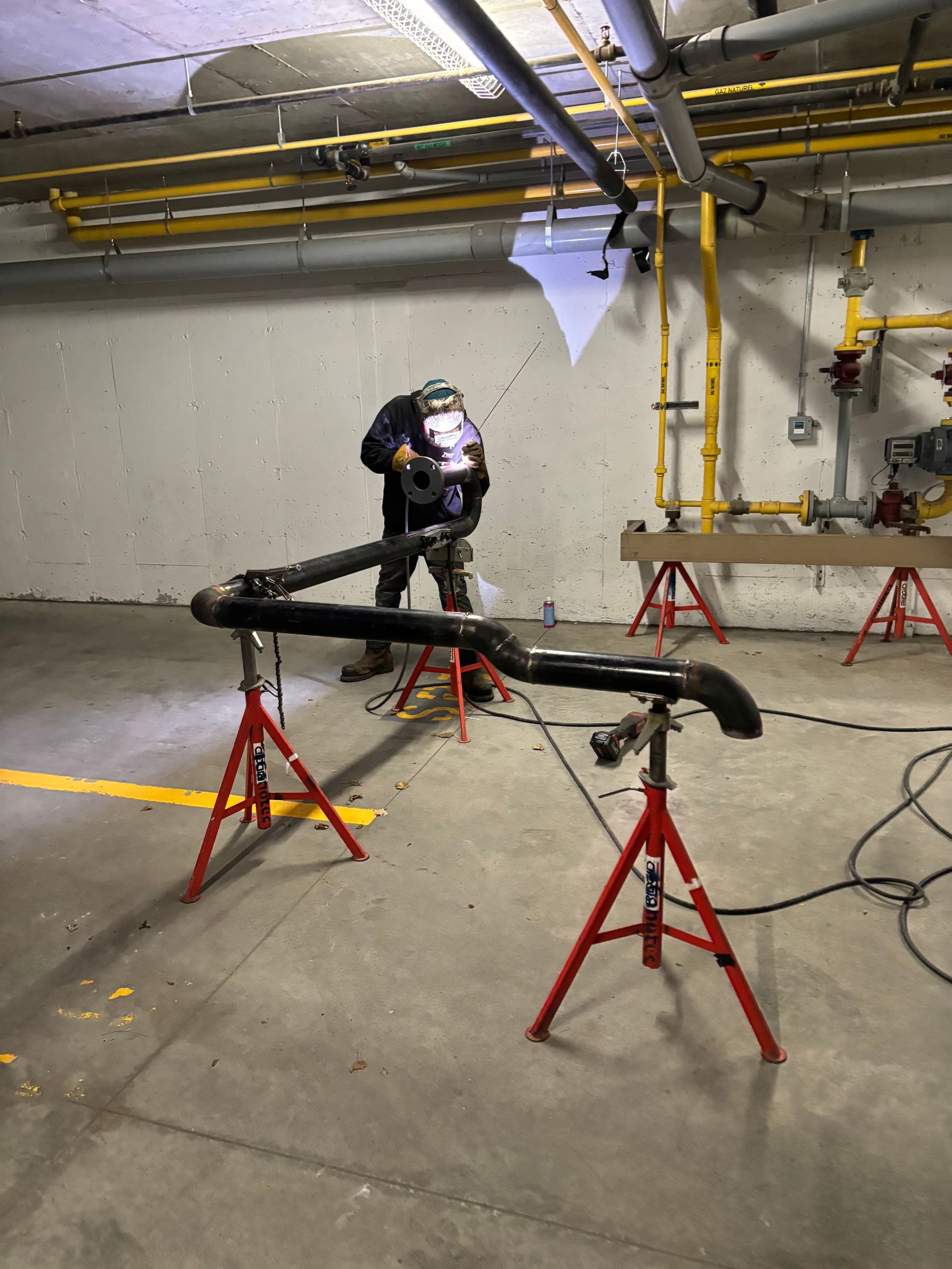 Welder welding pipe in a concrete room, wearing a mask. Black pipe on red stands.