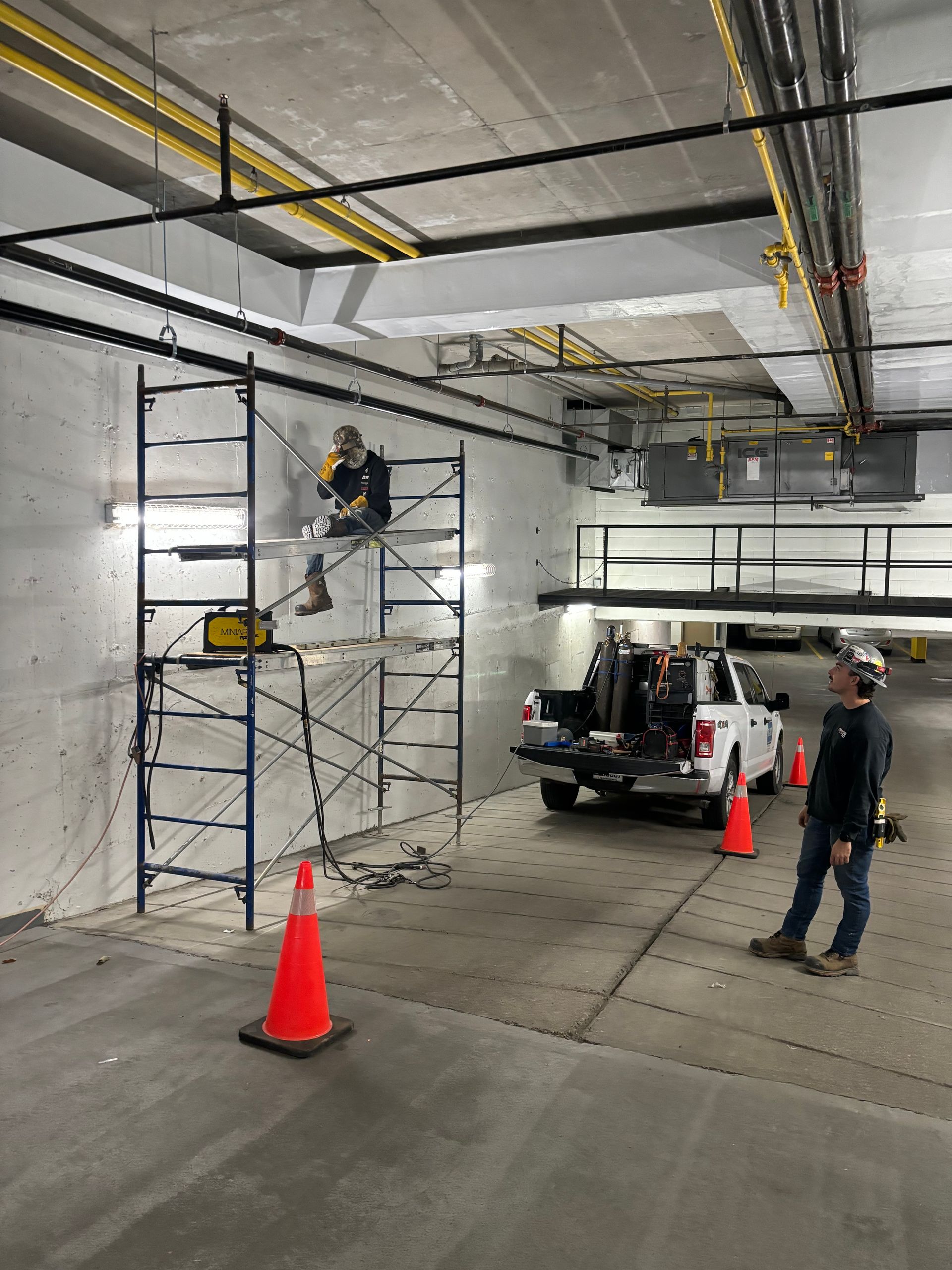 Workers in a parking garage: one on scaffolding, others near a truck. Orange cones, gray walls, and yellow pipes.