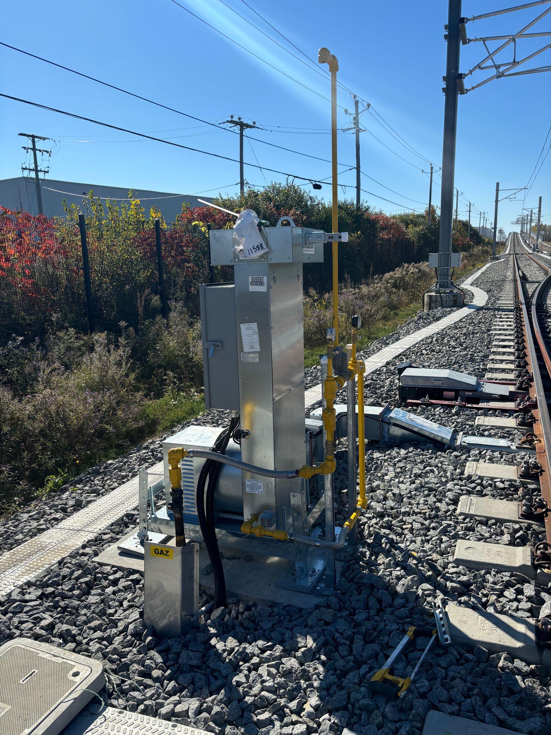 Railway signal equipment next to tracks, with overhead power lines and a sunny, outdoor setting.