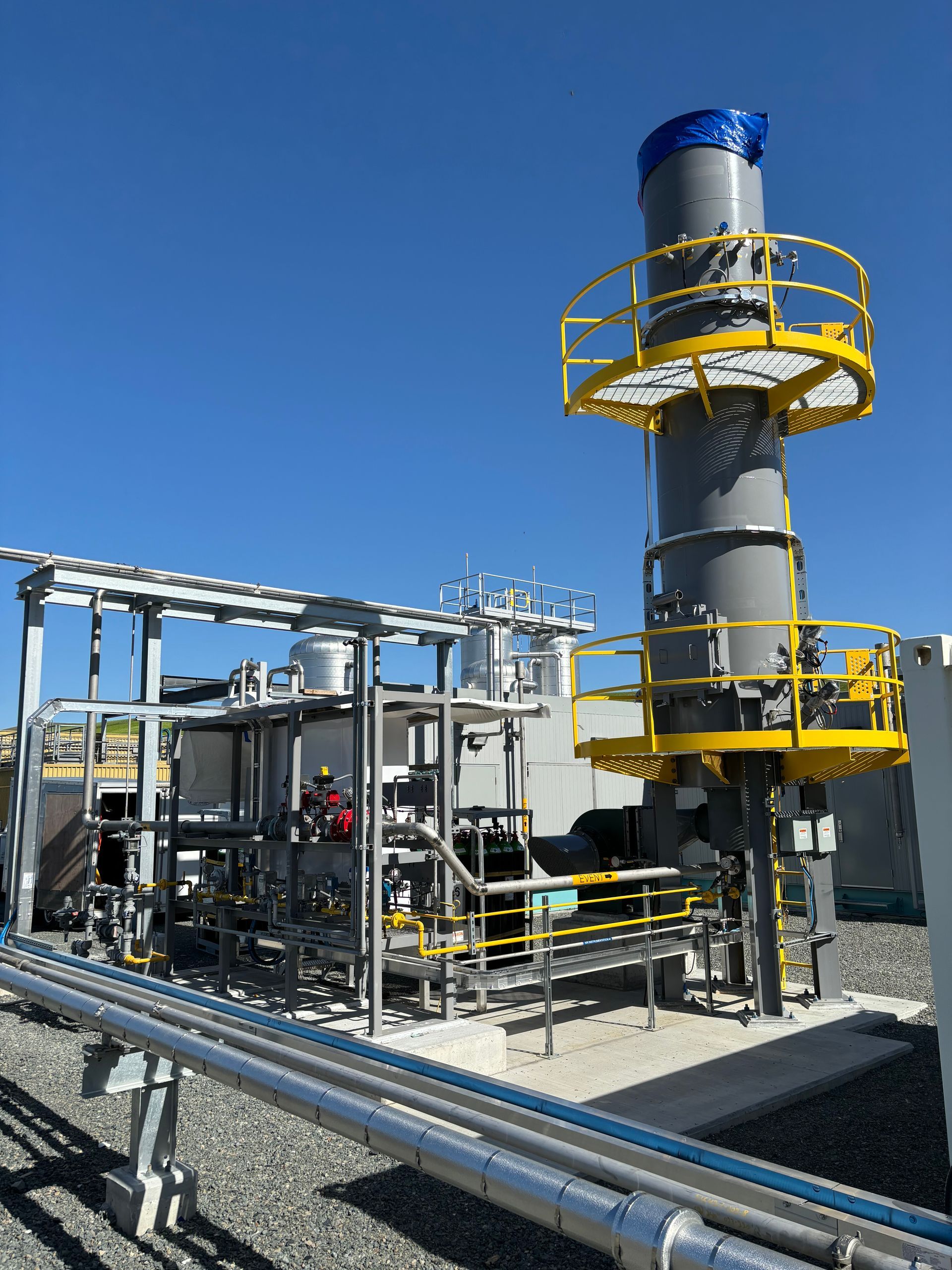 Industrial equipment with a tall, grey cylindrical stack and yellow scaffolding, under a blue sky.