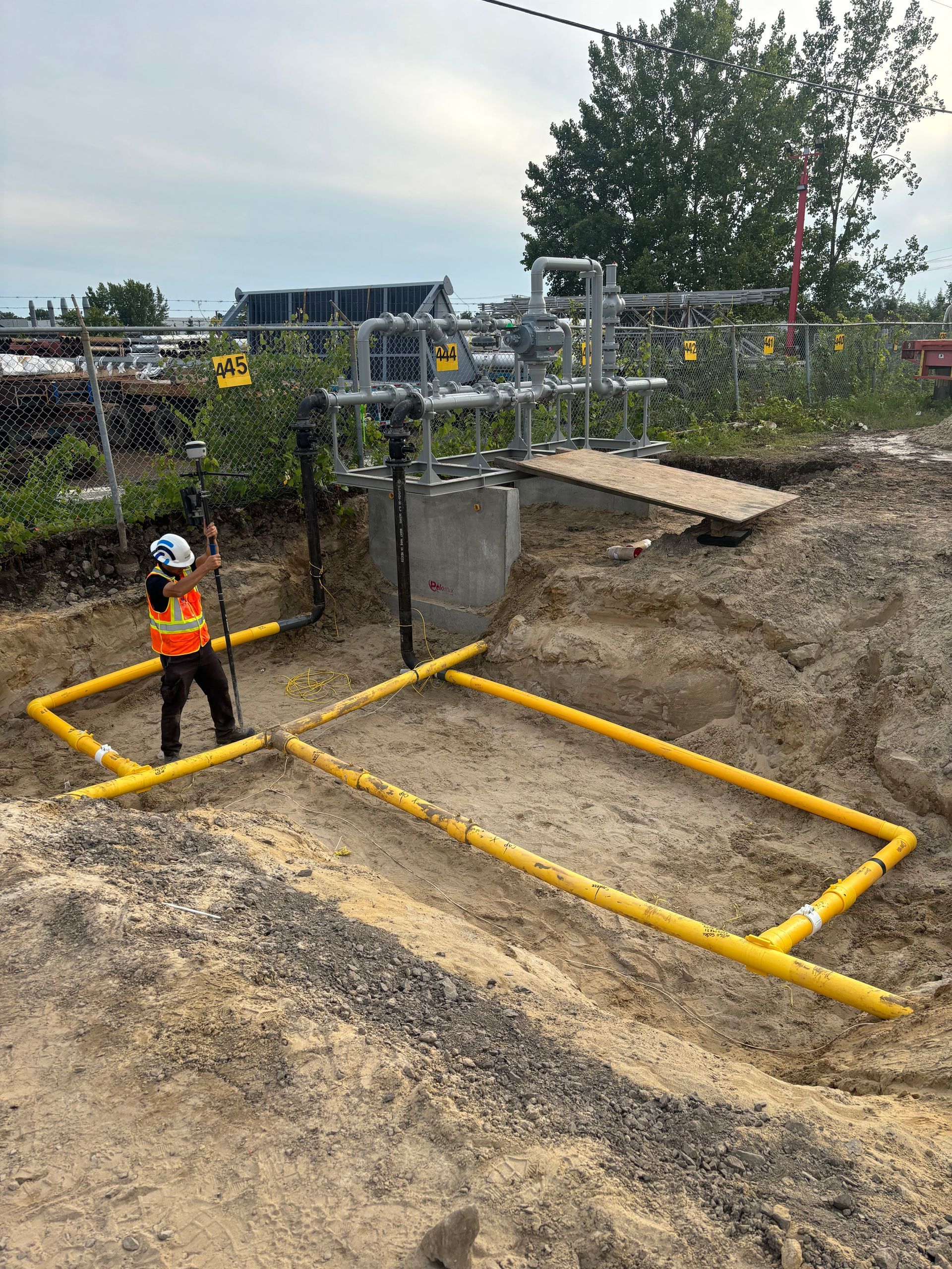 Construction worker near yellow gas pipes in a dirt pit; equipment overhead, outdoor setting.