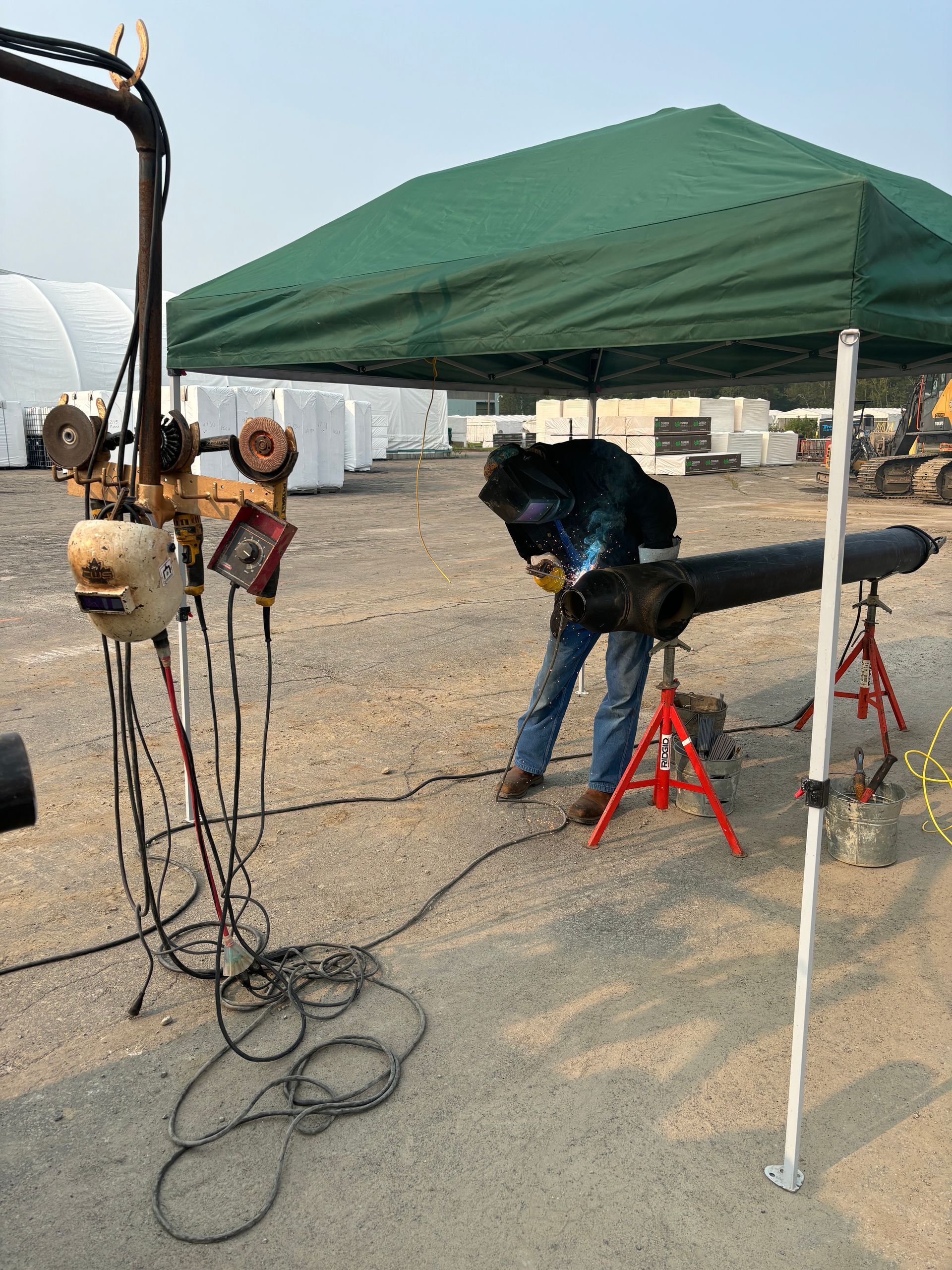 Welder in protective gear welding a pipe under a green canopy outdoors.