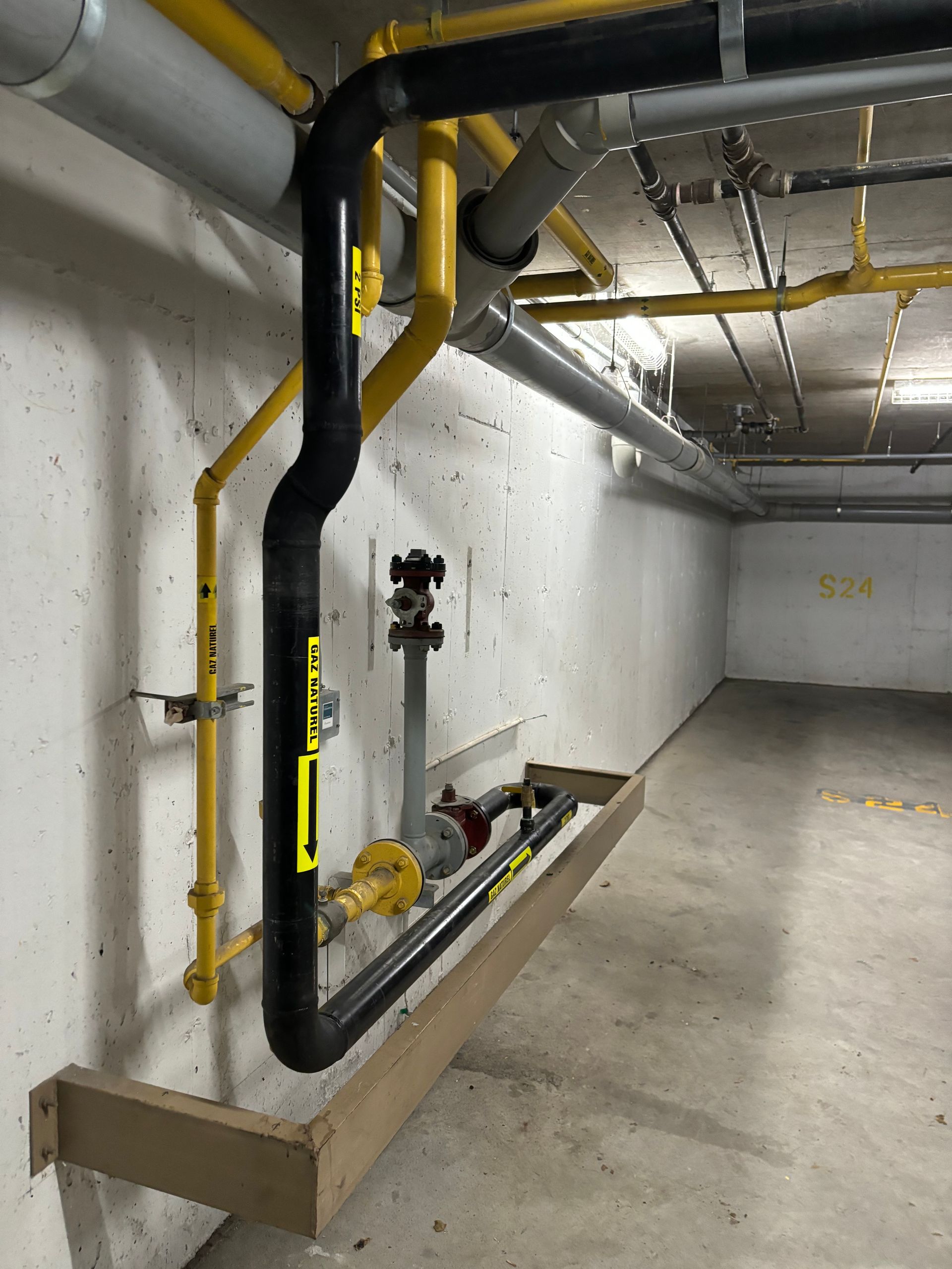 Pipes on a concrete wall. Black and yellow pipes run horizontally and vertically. A wooden beam is at the bottom.