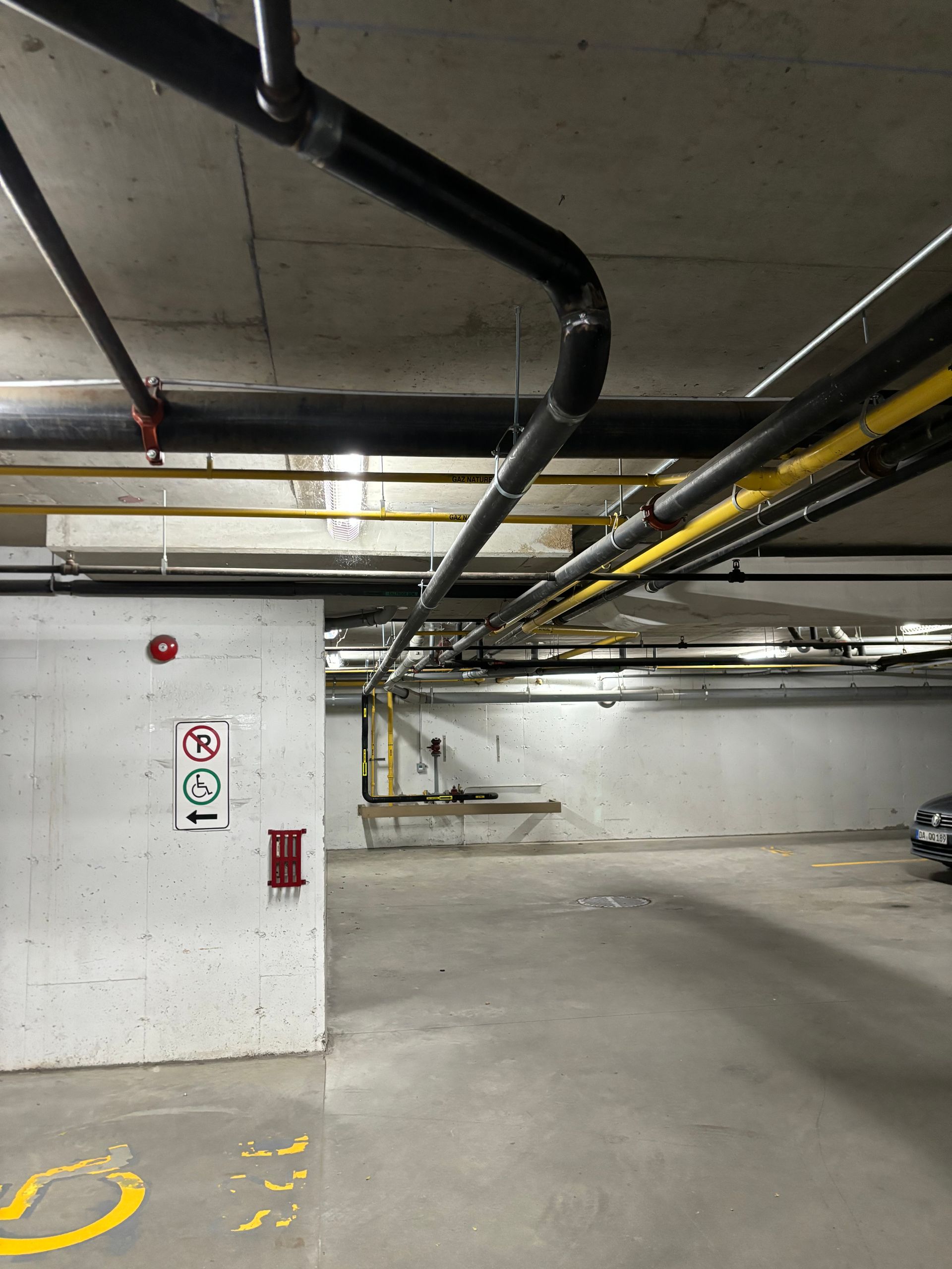 Parking garage with concrete walls and ceiling, pipes overhead. Fire safety equipment on the left.