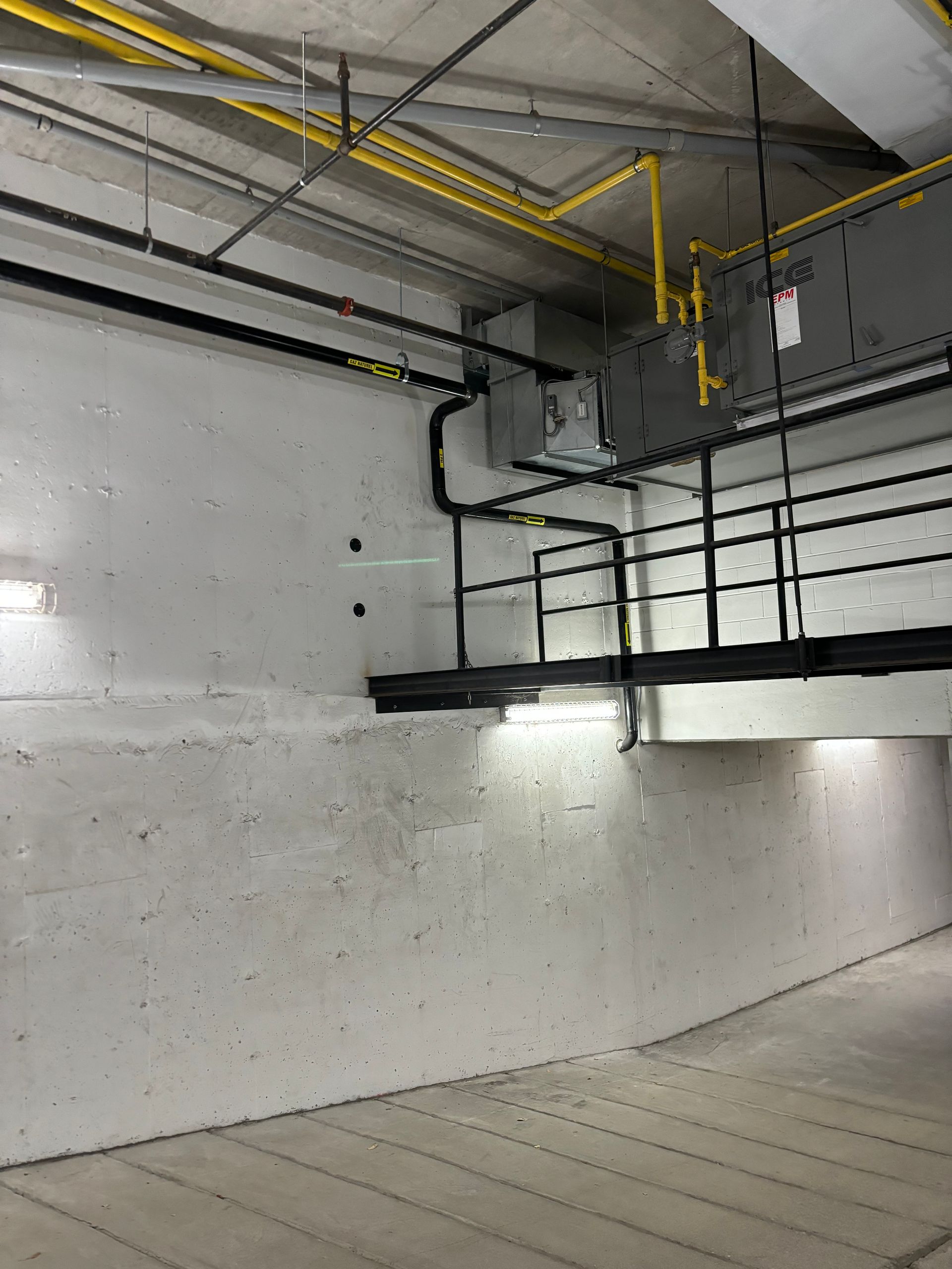 Concrete wall in a parking garage with black piping, a metal platform, and yellow gas lines overhead.
