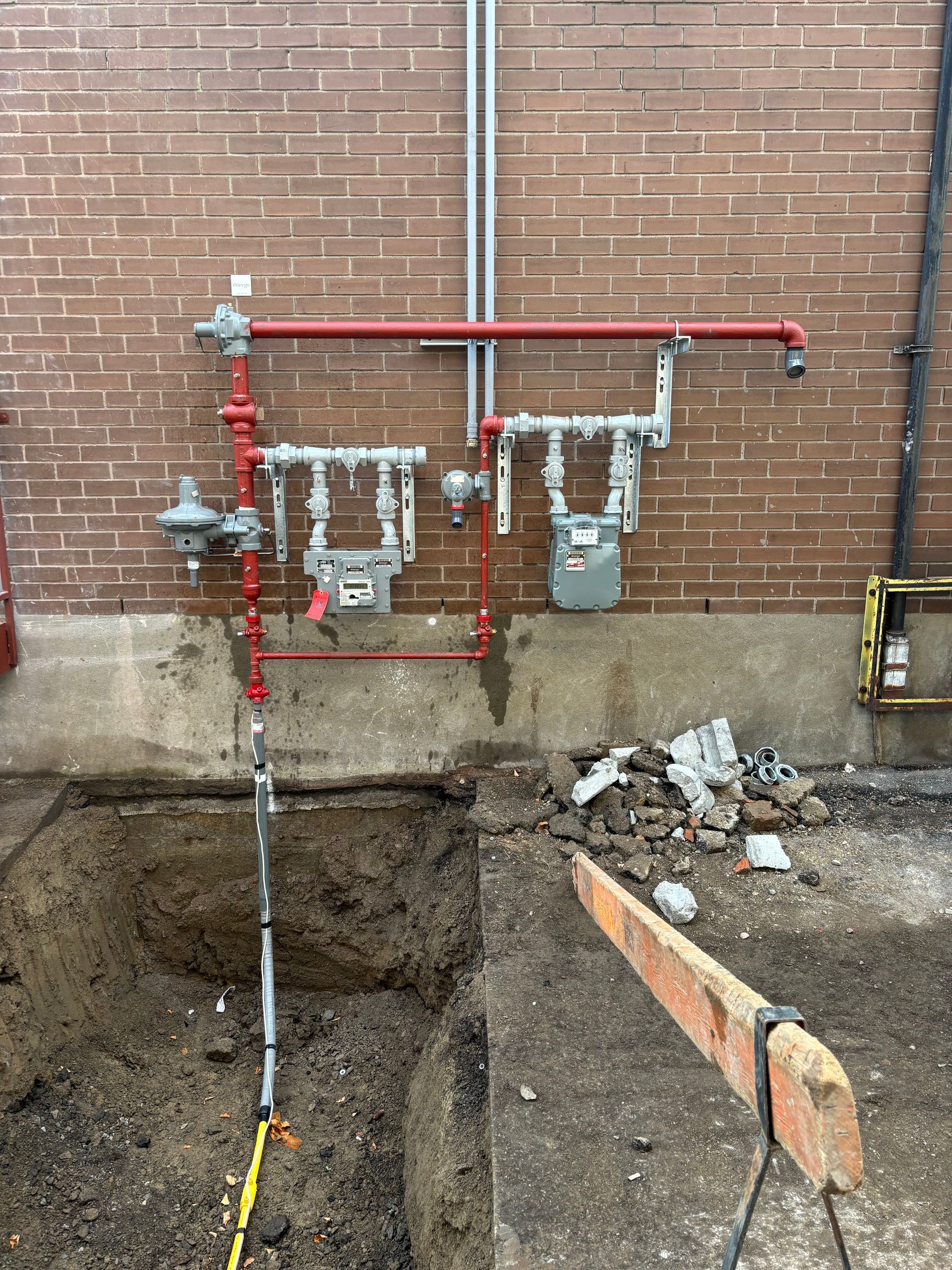 Gas meters and fire sprinkler system on brick wall, with excavated ground in front.