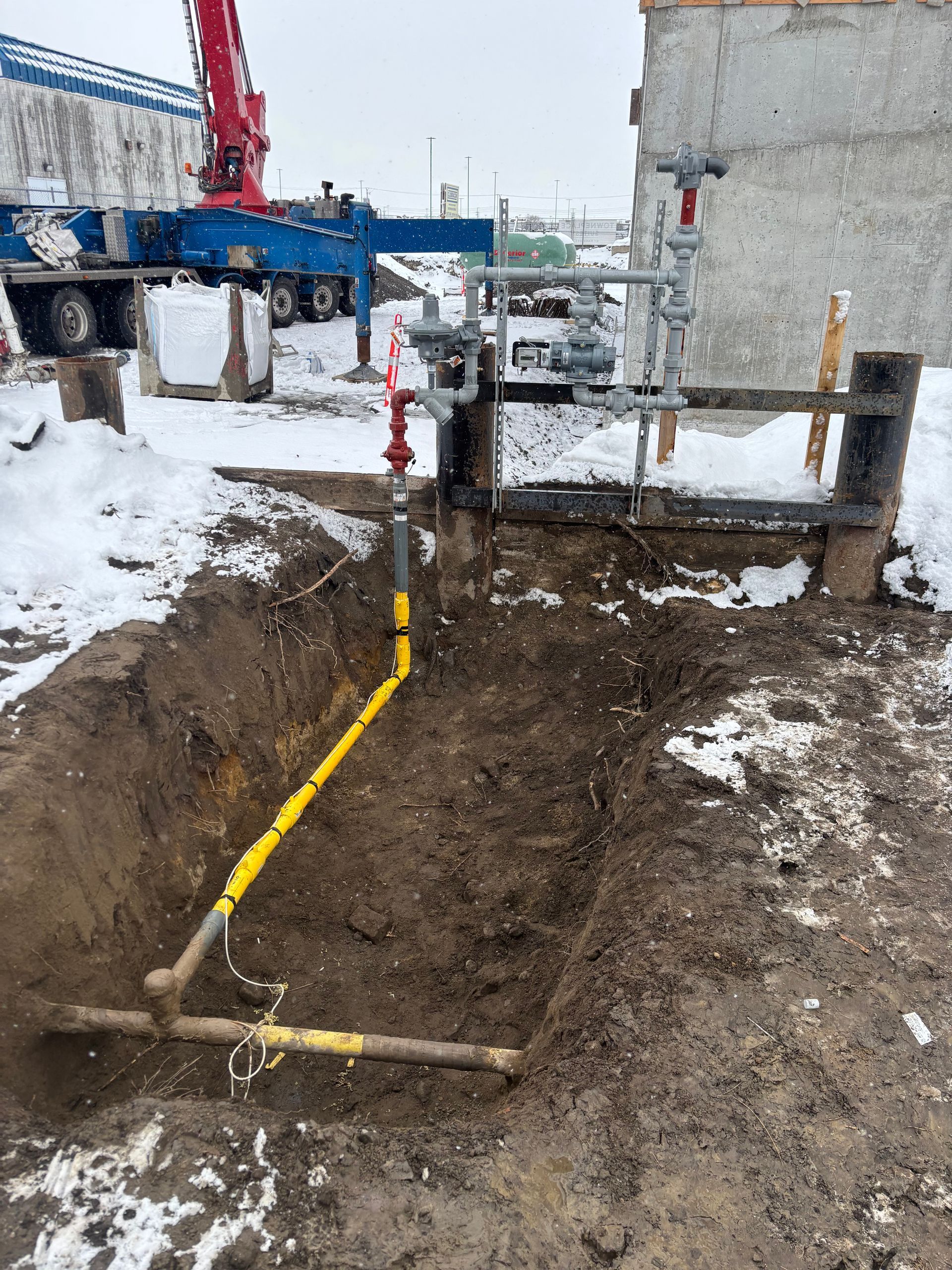 A yellow gas line in an open trench in the snow near a construction site.