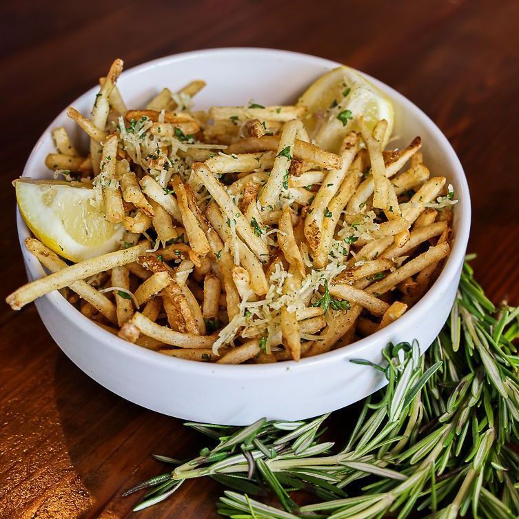 A bowl of french fries with lemon and rosemary on a wooden table.
