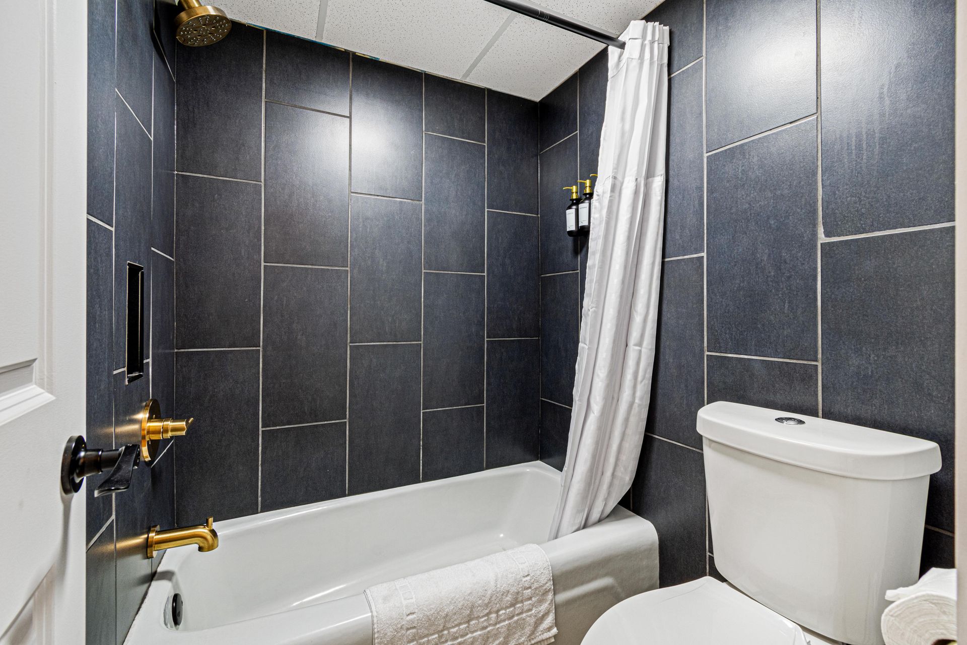 Dark gray tiled bathroom with gold fixtures, white tub, toilet, and shower curtain.