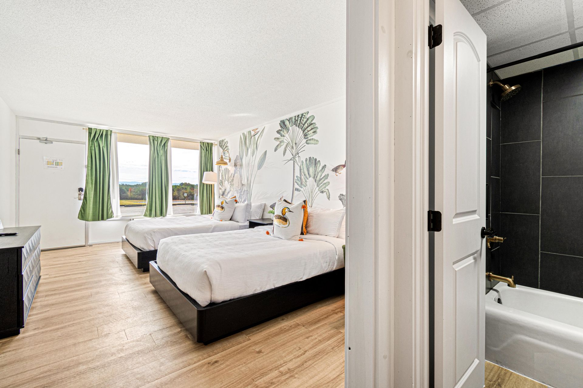 Hotel room with two beds, floral wall, green curtains, and bathroom visible.