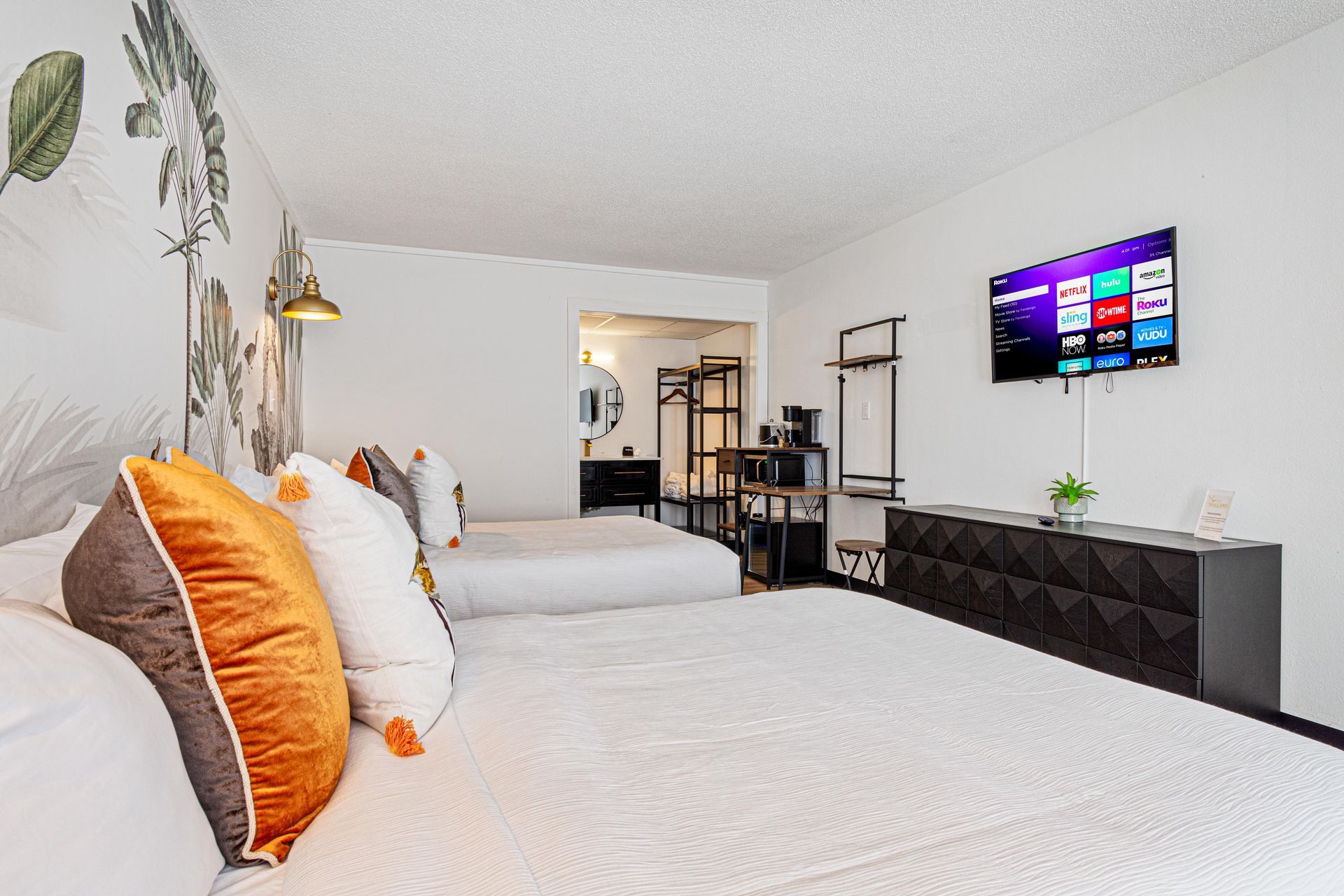 Two queen beds in a bright hotel room with decorative pillows, TV, and a small kitchenette.