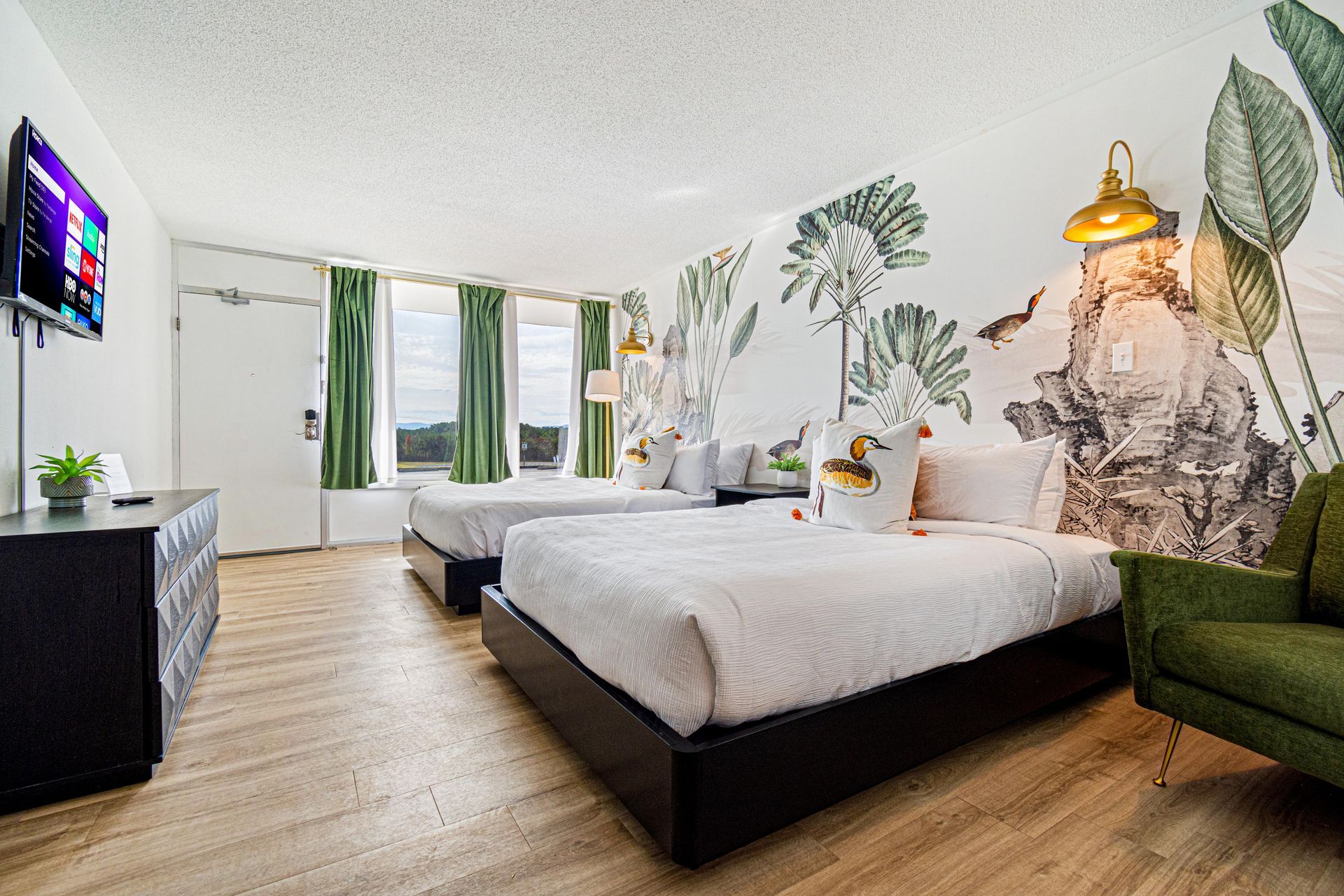 Hotel room with two beds, green couch, TV, and a wall mural featuring tropical plants.