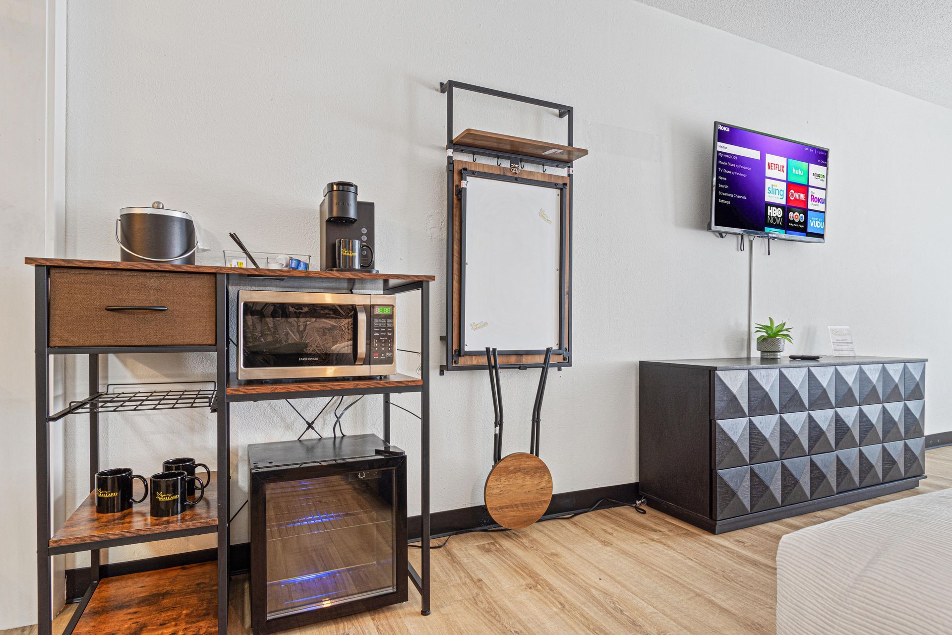 Room with a microwave, coffee maker, mini fridge, and TV. Wooden floors, black and brown furniture, white walls.